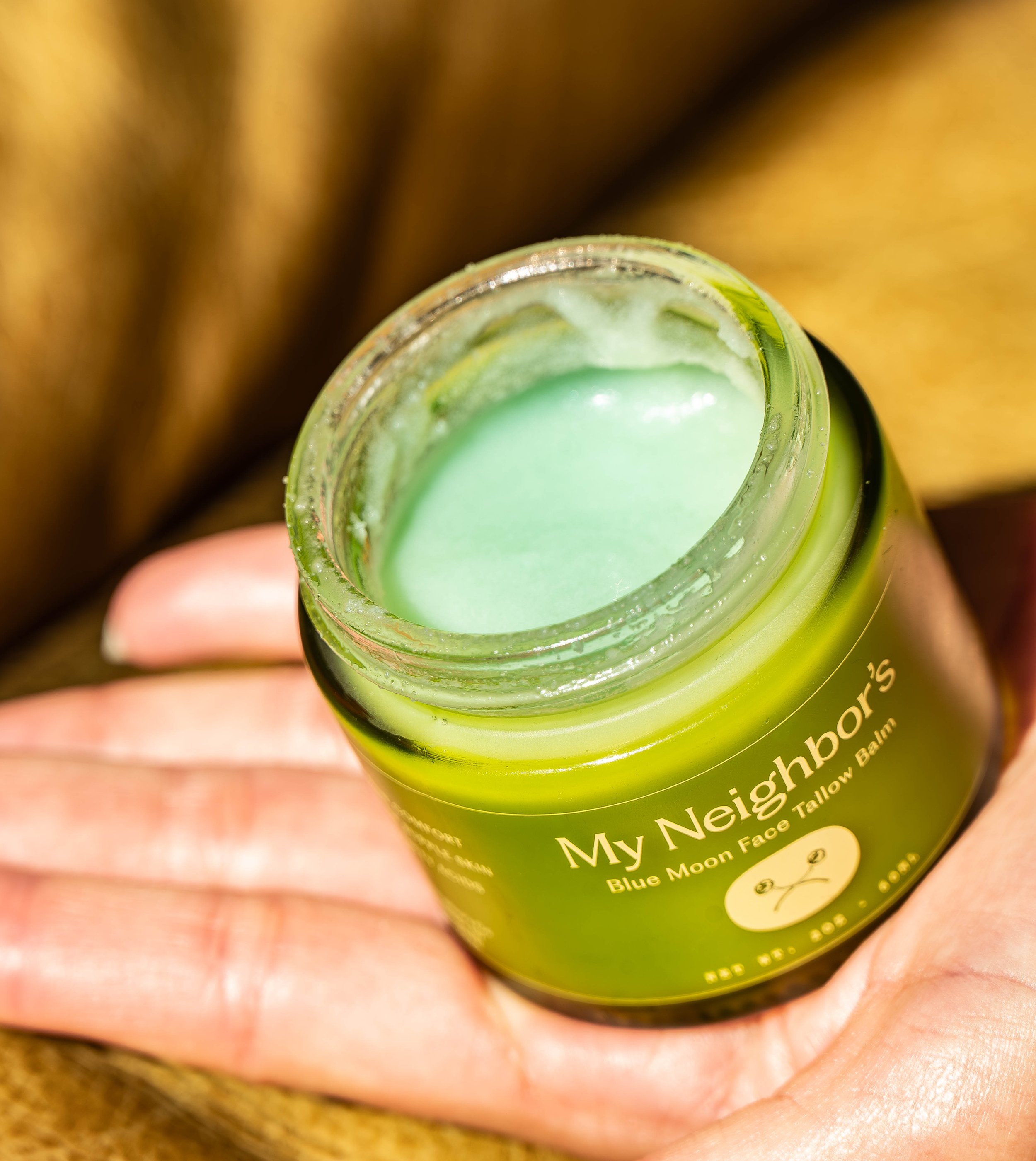 A small jar of green cream or balm labeled "My Neighbours Moon Face Tallow Balm" being held in a person's hand.