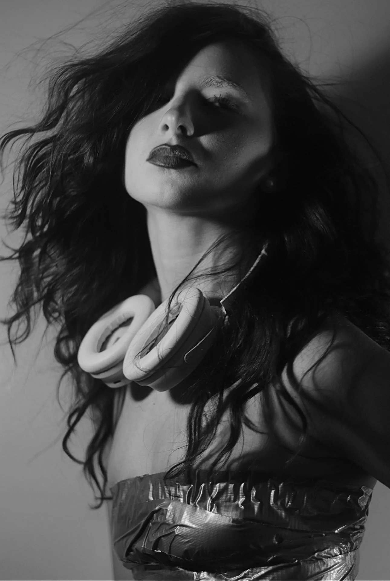 A woman with dark, wavy hair wears headphones around her neck and a strapless top. The image is in black and white.