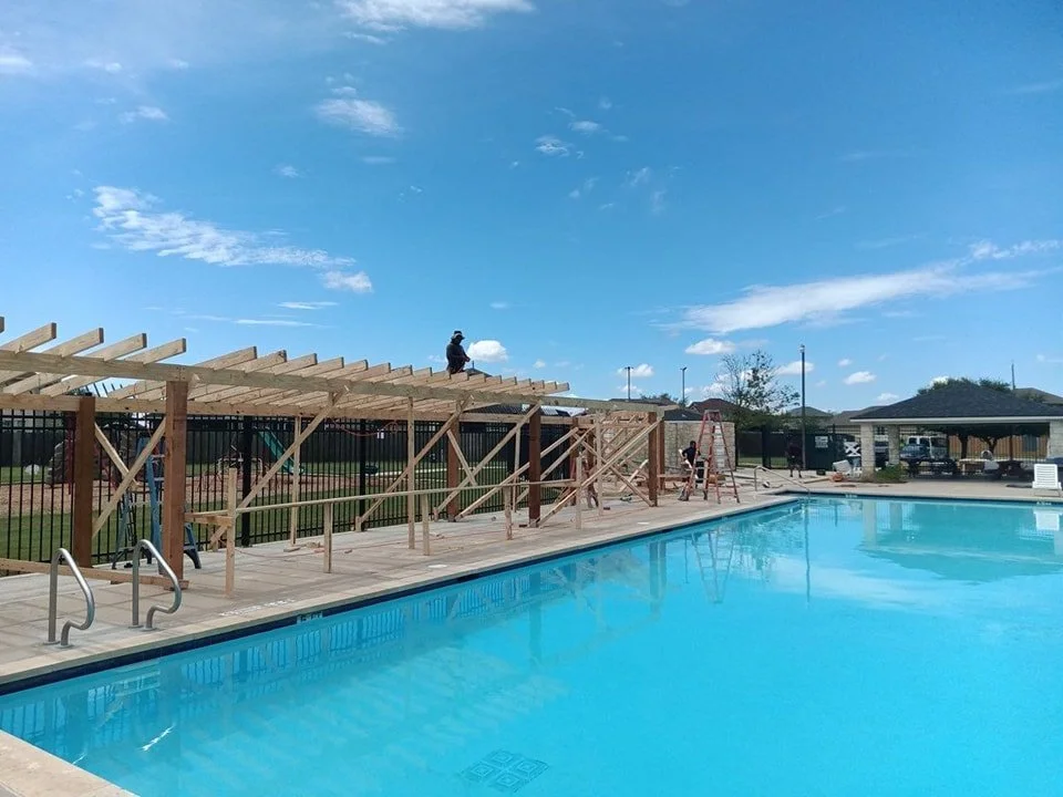 Construction workers building a wooden structure near an outdoor swimming pool on a sunny day.