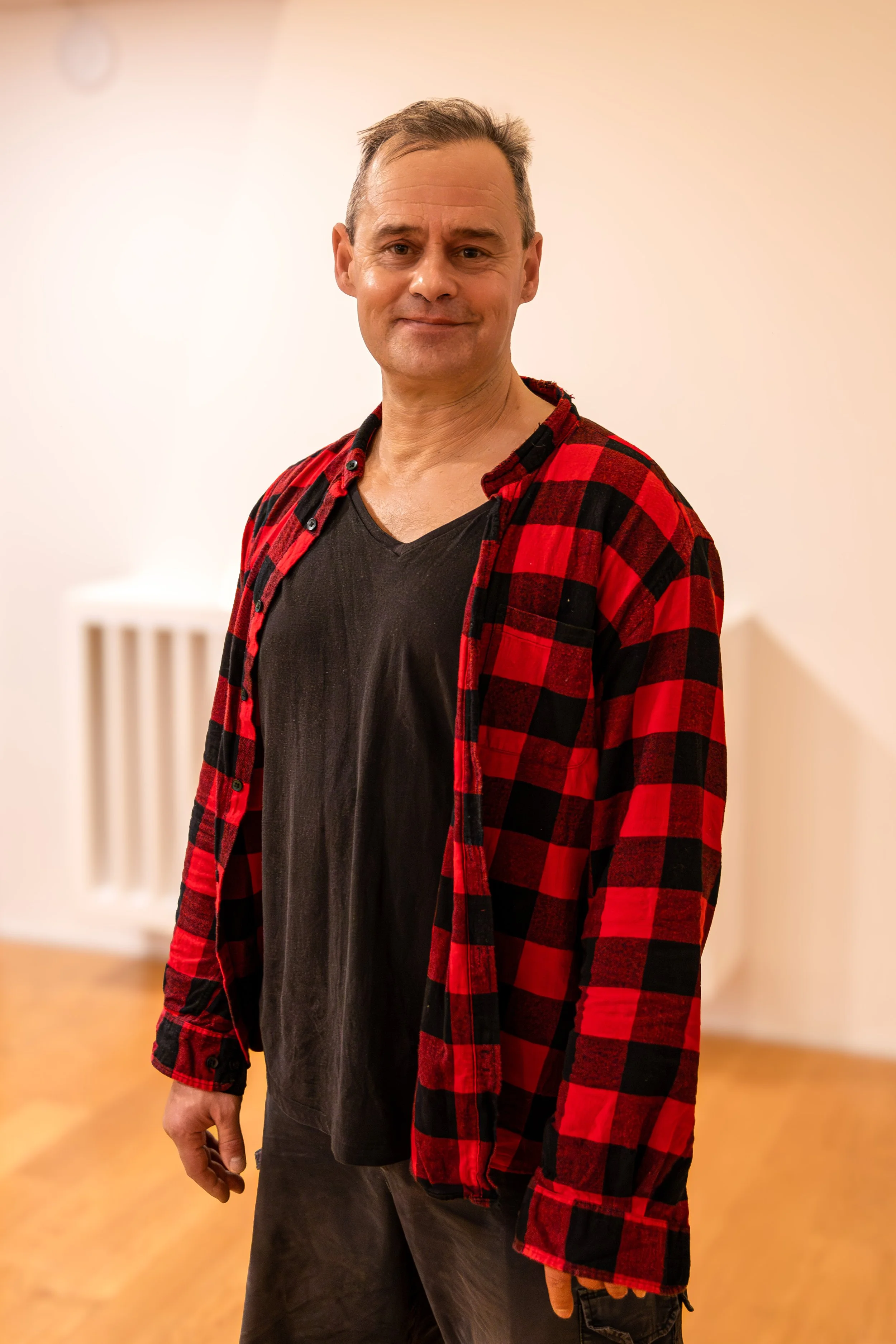 Julien Diaz wearing a red and black plaid shirt over a black t-shirt, standing indoors on a wooden floor.