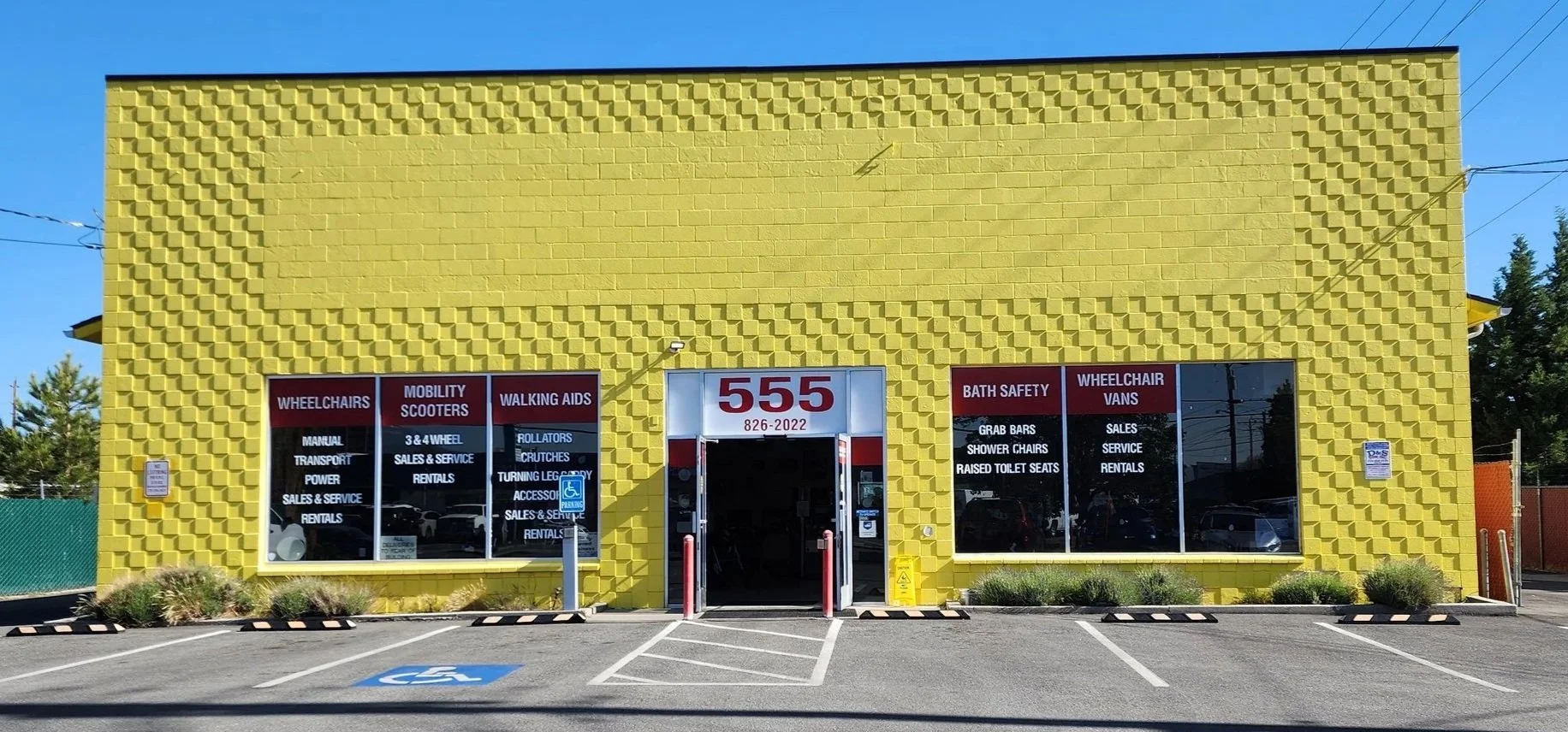 A yellow building with large windows displaying various mobility products like wheelchairs, scooters, walkers, and bath safety items. The entrance has the number 555 and a date, with a parking lot in front marked for handicapped parking.