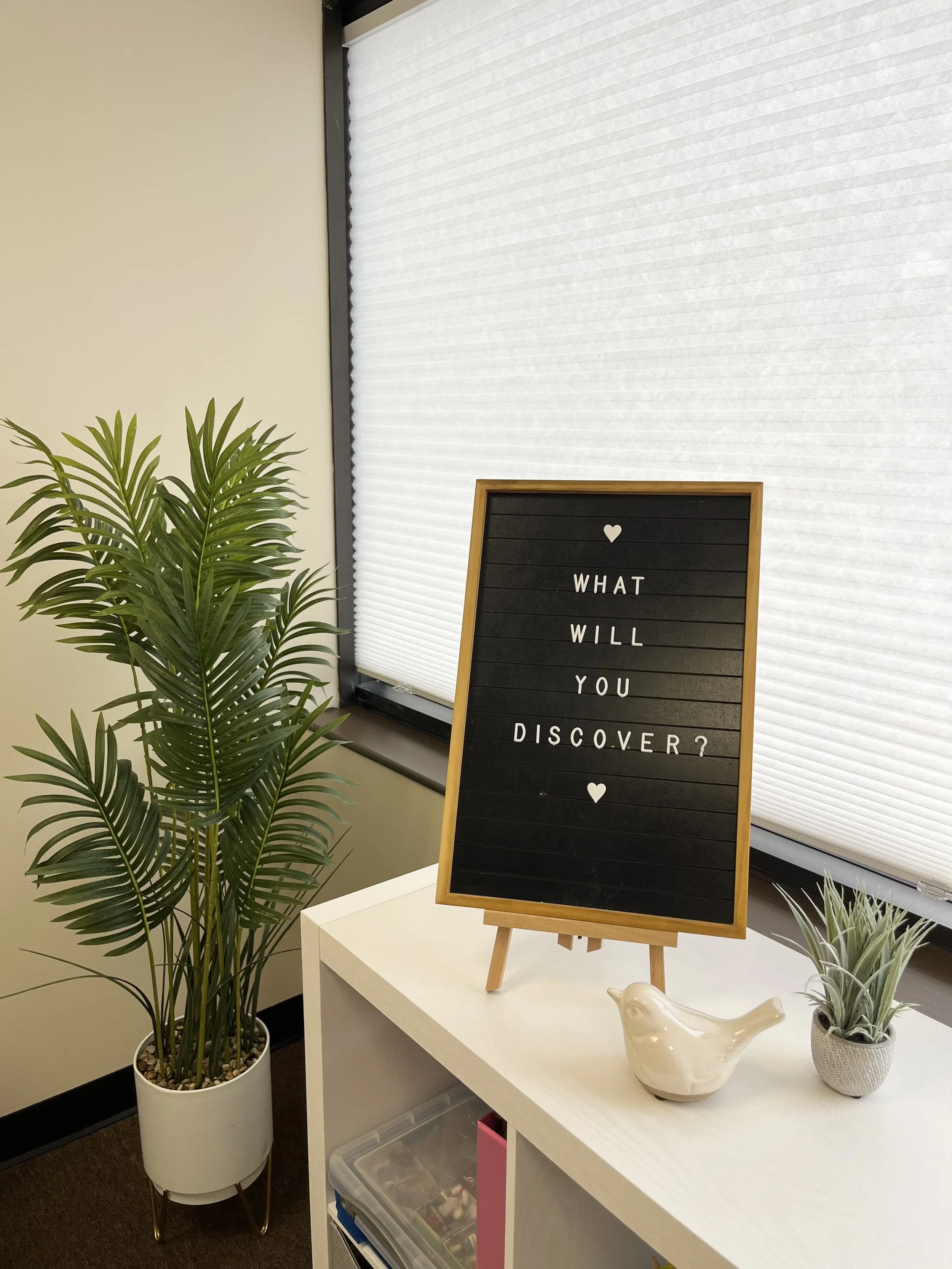 A black letter board with the message 'What will you discover?' flanked by a white ceramic bird figurine and a small potted plant, placed on a white table next to a window with closed white blinds and a tall potted palm plant.