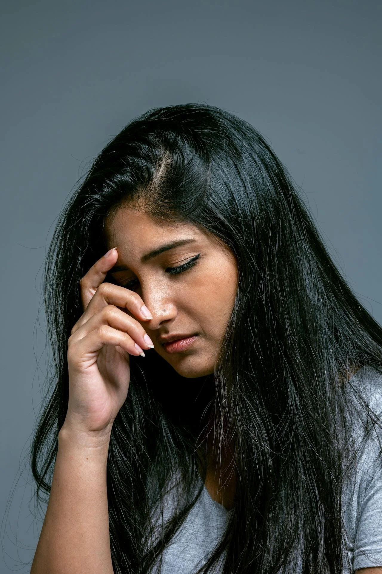 A woman with long black hair and a small nose piercing holding her forehead with her eyes closed, appearing stressed or worried.