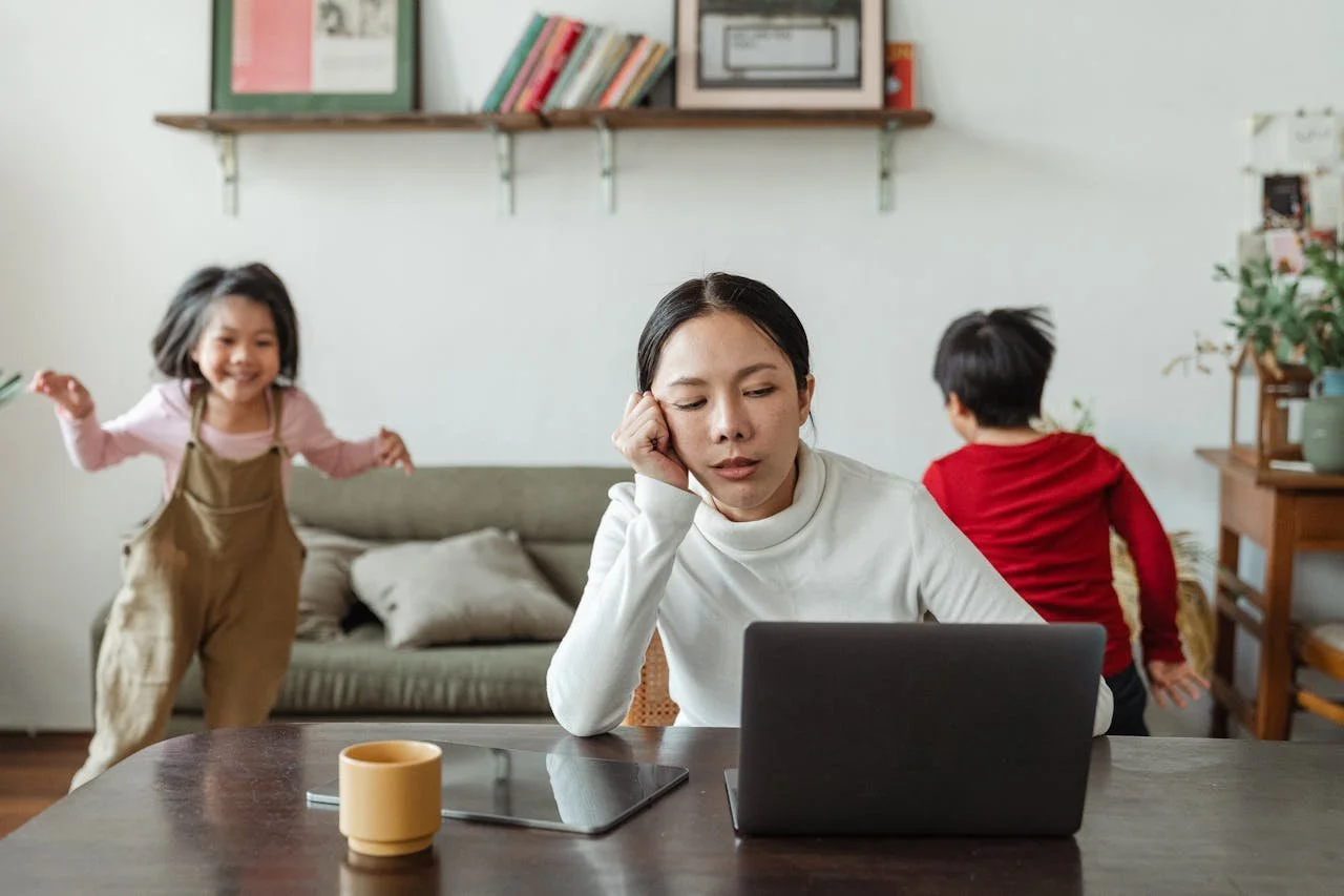 An woman with dark hair sitting at a desk in front of a laptop, looking tired or bored, with two children playing in the background in a cozy home setting.
