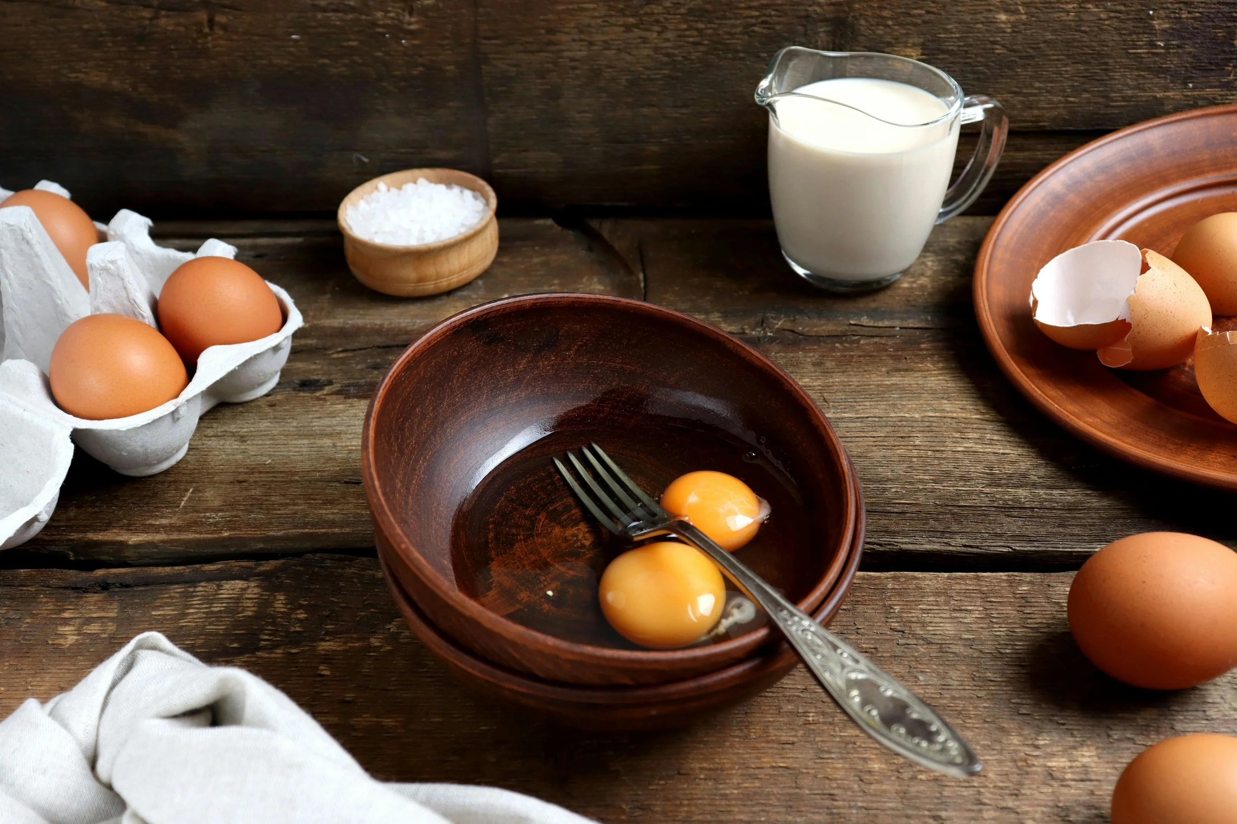 A wooden bowl with two eggs and a fork with a carton of eggs and bowl of salt beside it