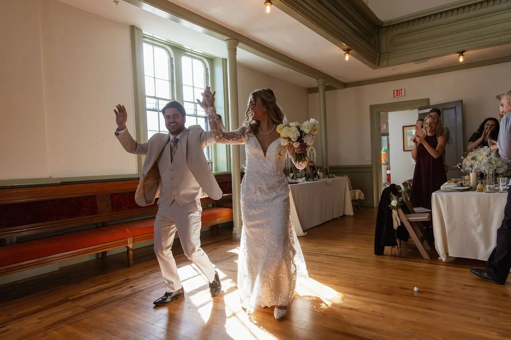A bride and groom dancing at their wedding reception, surrounded by guests in a decorated indoor venue with wooden floors and large windows.