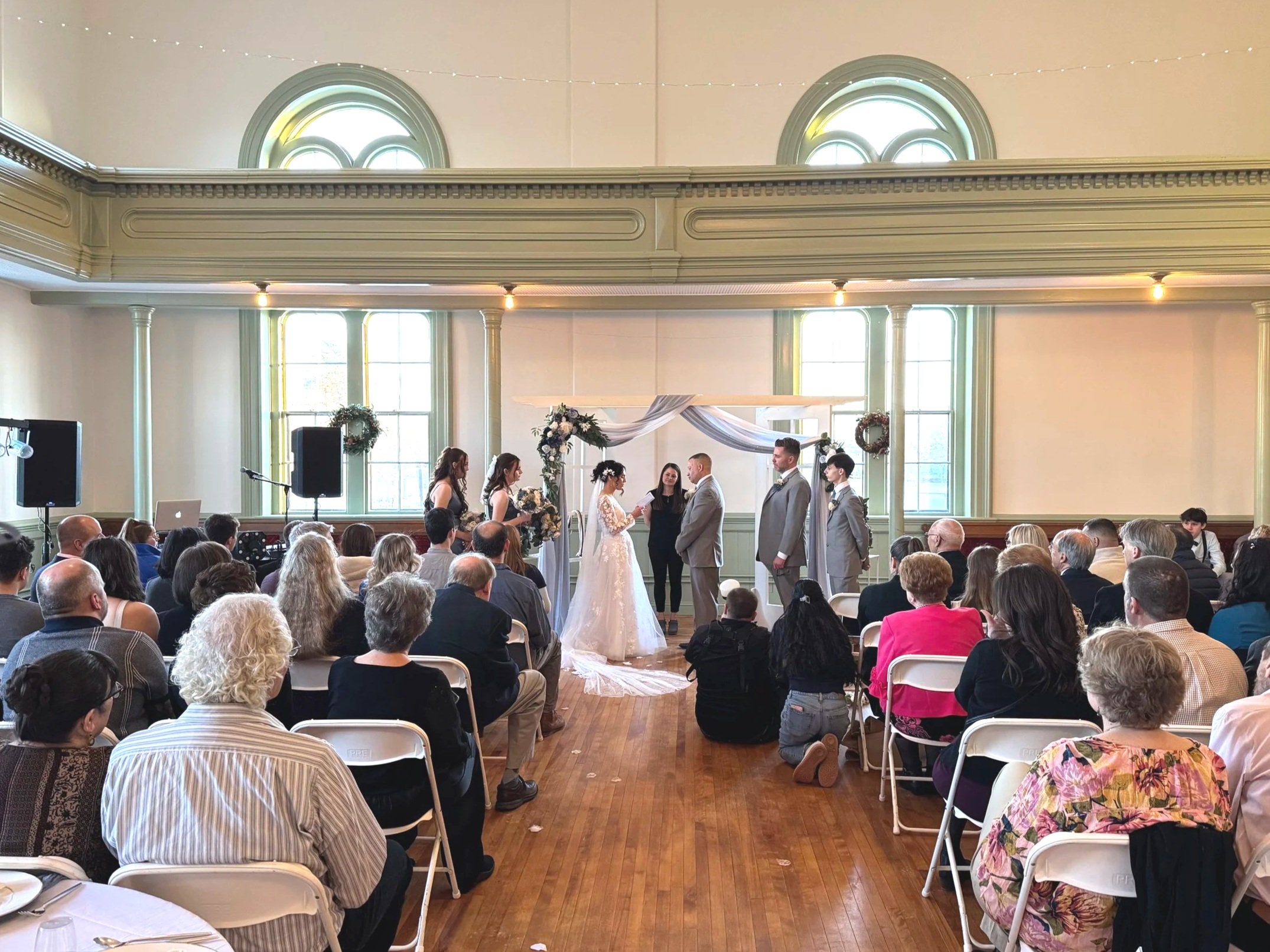 A wedding ceremony taking place in a spacious indoor venue with high ceilings and large windows. The bride and groom stand in front of an officiant, surrounded by bridesmaids and groomsmen, with seated guests observing the event.