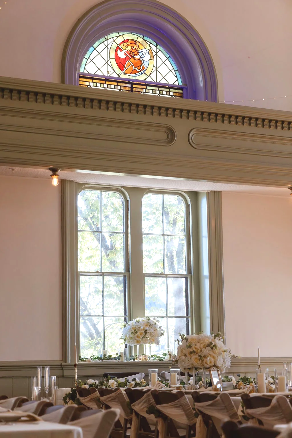 Bright room with large window and white floral centerpieces on decorated tables, stained glass window on upper wall, and light fixtures
