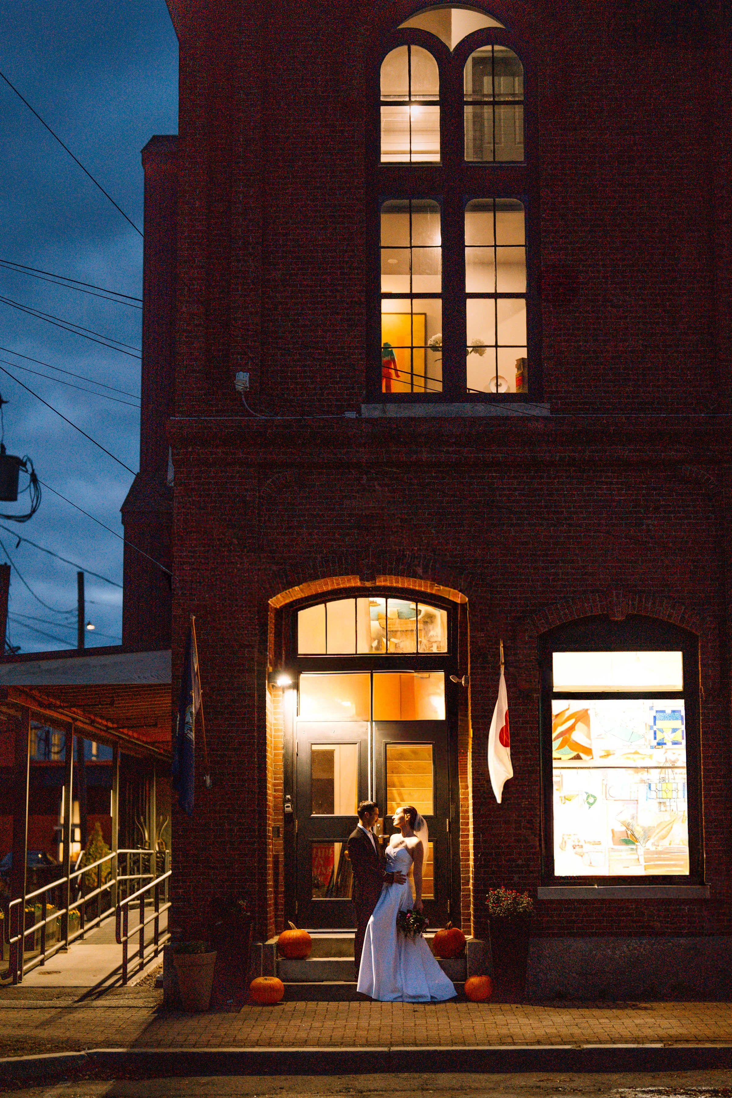 A newly married couple in wedding attire stands outside a lit building at night, sharing a kiss near the entrance decorated with pumpkins.
