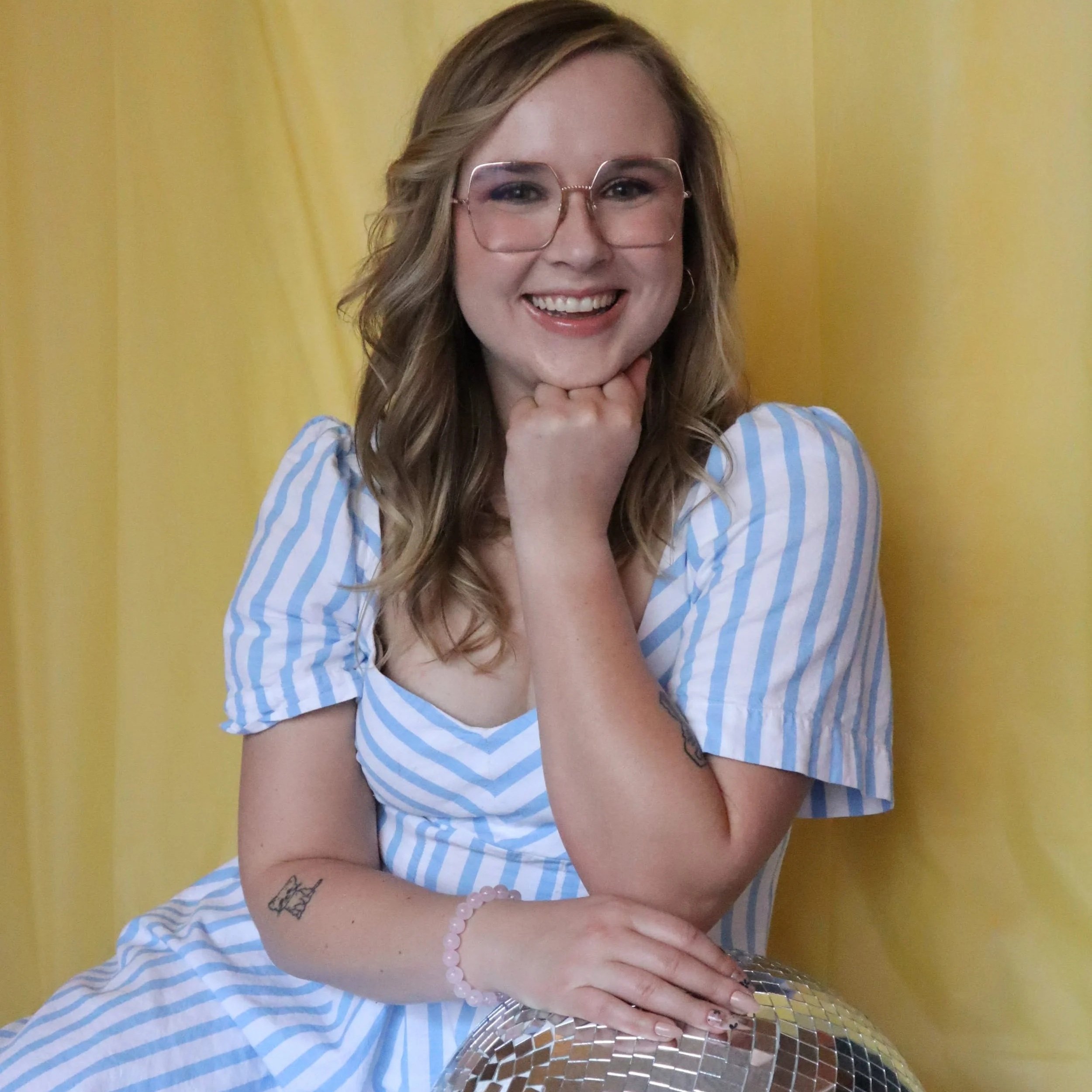 Moriah, the owner of JayBird Events, poses in front of a yellow backdrop. She is wearing a blue and white striped dress