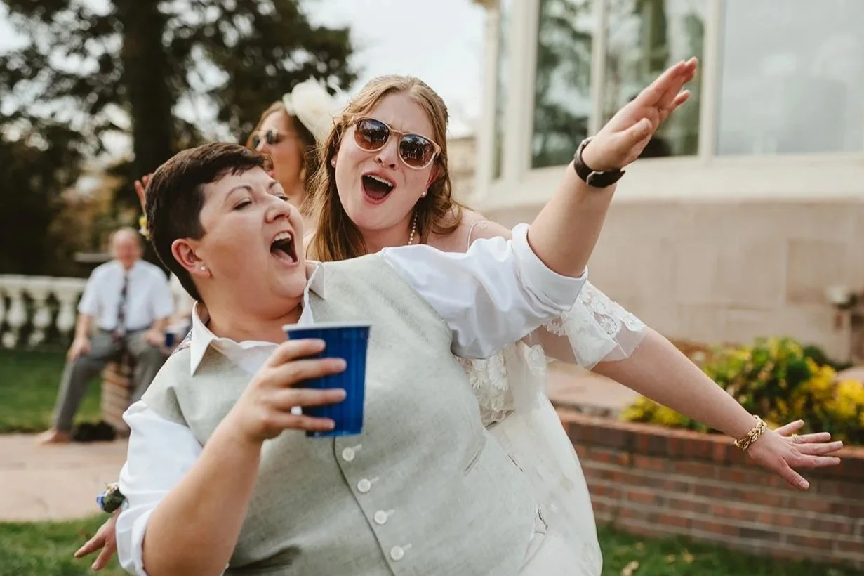 Two brides sing and dance together at their wedding planned by JayBird Weddings and Events. One of them is holding a drink.
