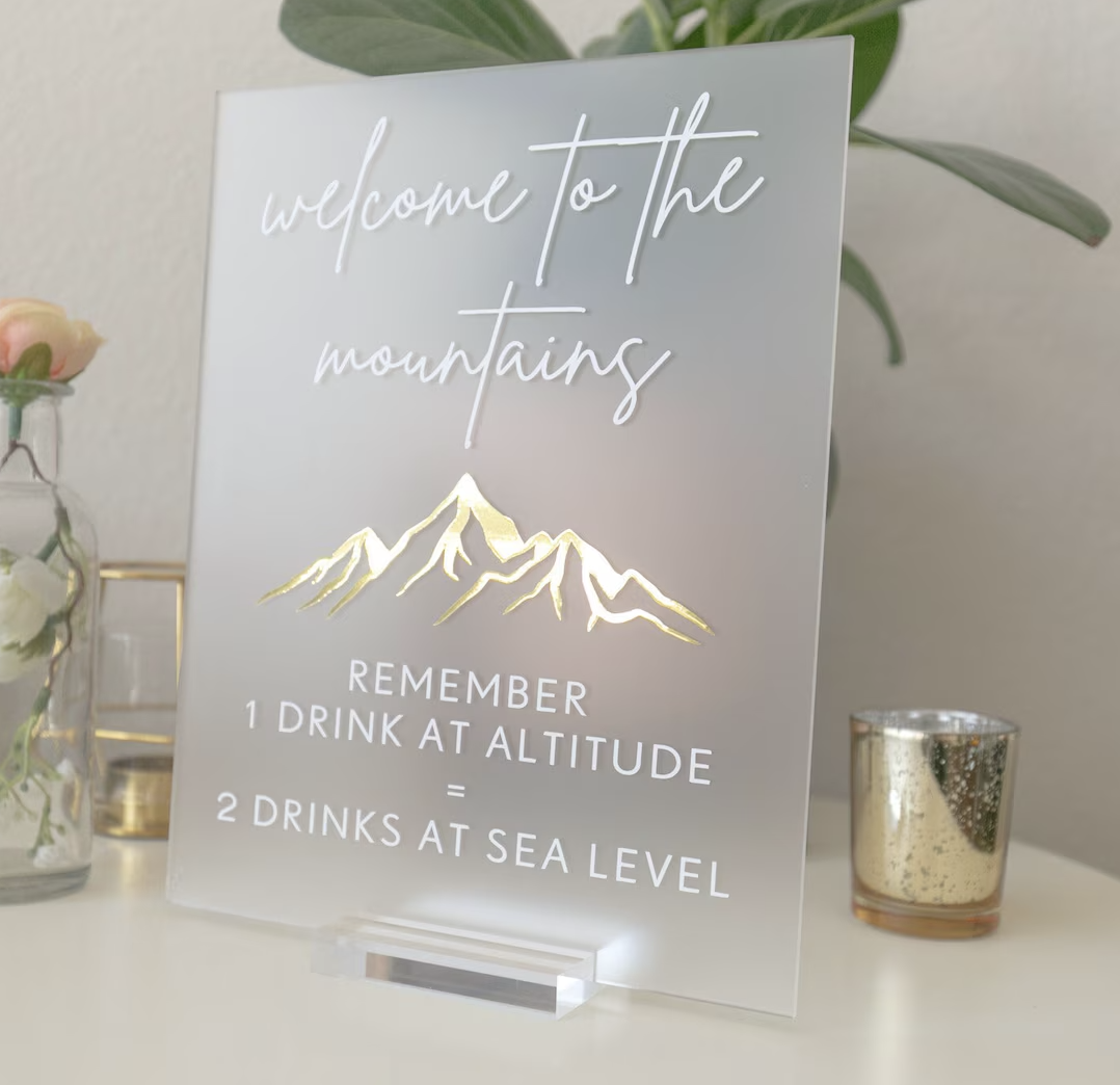 Acrylic sign with white letters that say "welcome to the mountains. Remember 1 drink at altitude = 2 drinks at sea level" from JayBird Weddings and Events