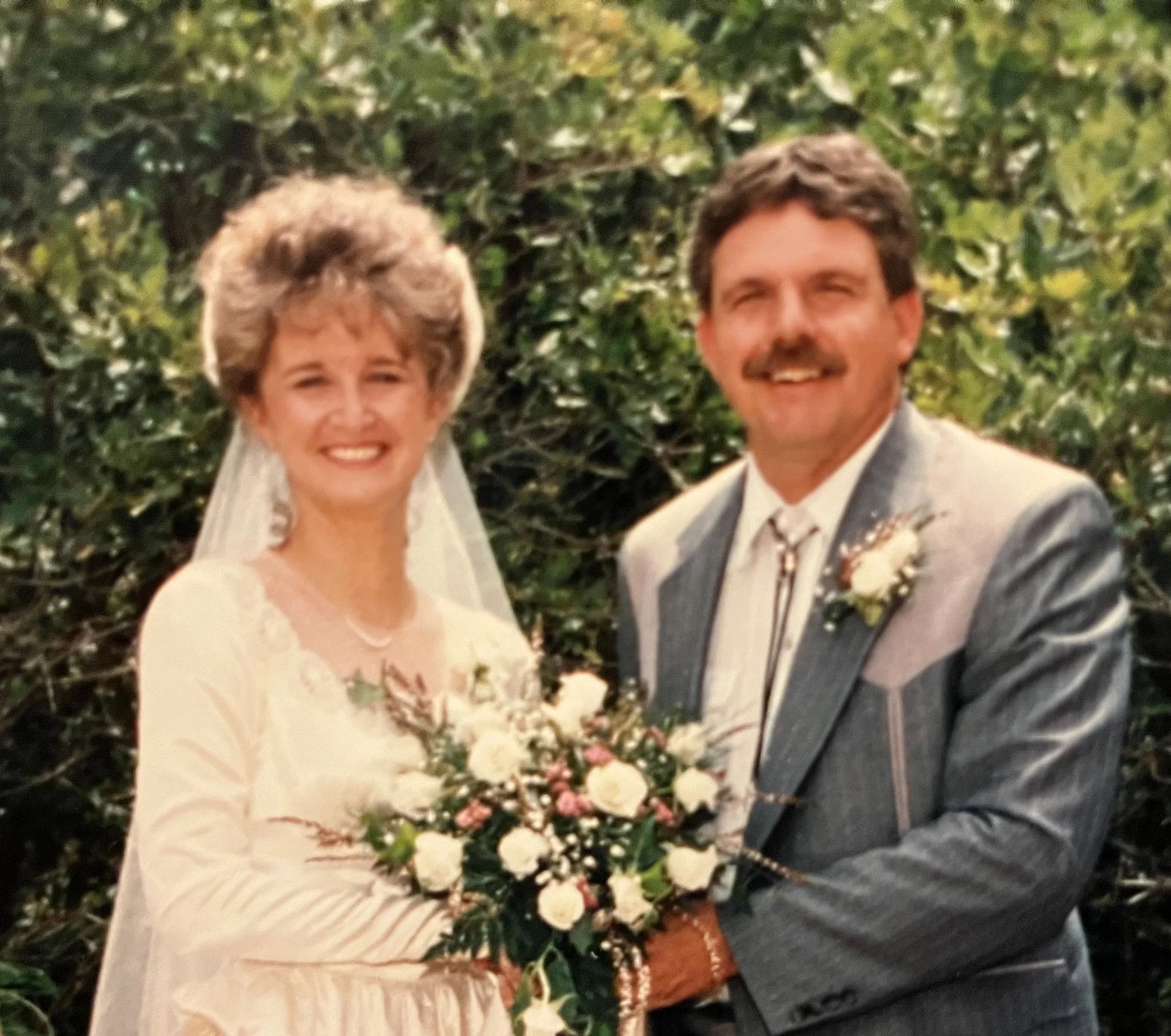 A couple poses in an old wedding photo from 1993. A bride and groom stand holding hands facing the camera in front of a green background of foliage