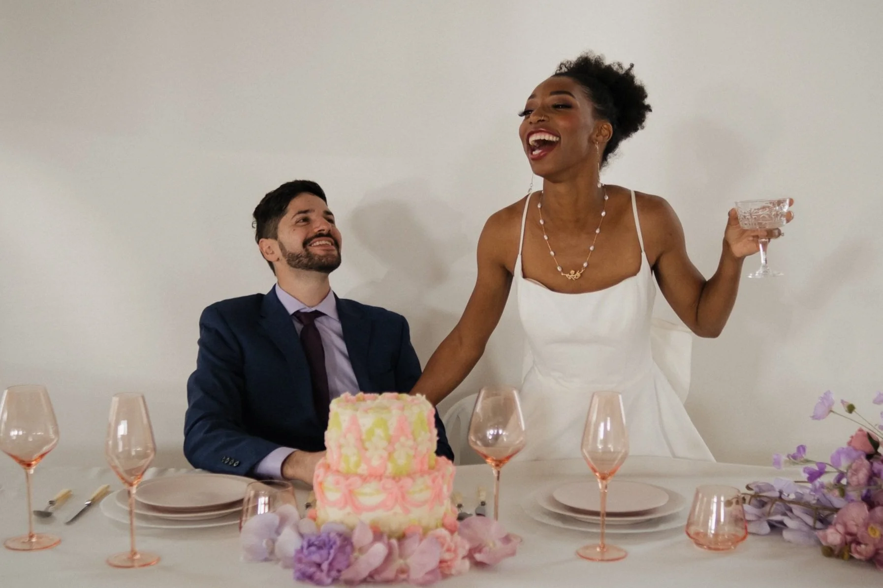A bride stands and laughs while holding a glass next to her groom, who is seated behind a colorful two-tiered cake, during a wedding planned by JayBird Weddings and Events.