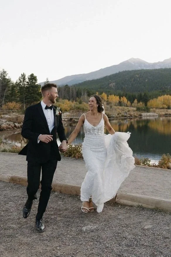 A bride and groom run hand-in-hand alongside a lake in the mountains of Colorado as they take a break from their wedding planned by JayBird Weddings and Events.