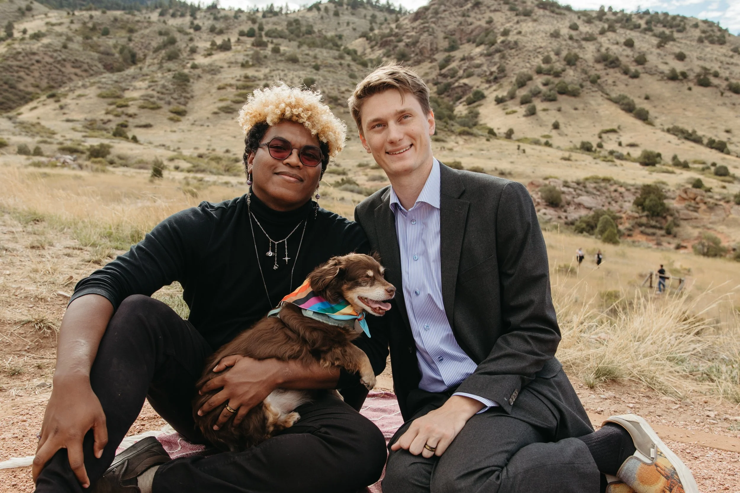 A gay couple sit together on a picnic blanket with their dog, who is wearing a multicolor bandana, during their wedding that was planned by JayBird Weddings and Events.