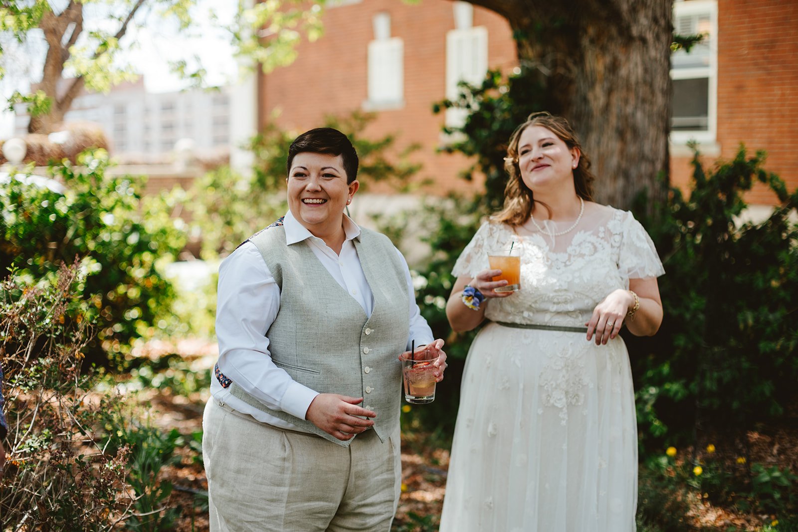 Two brides smile and enjoy cocktails on their wedding day, outside in Denver, Colorado