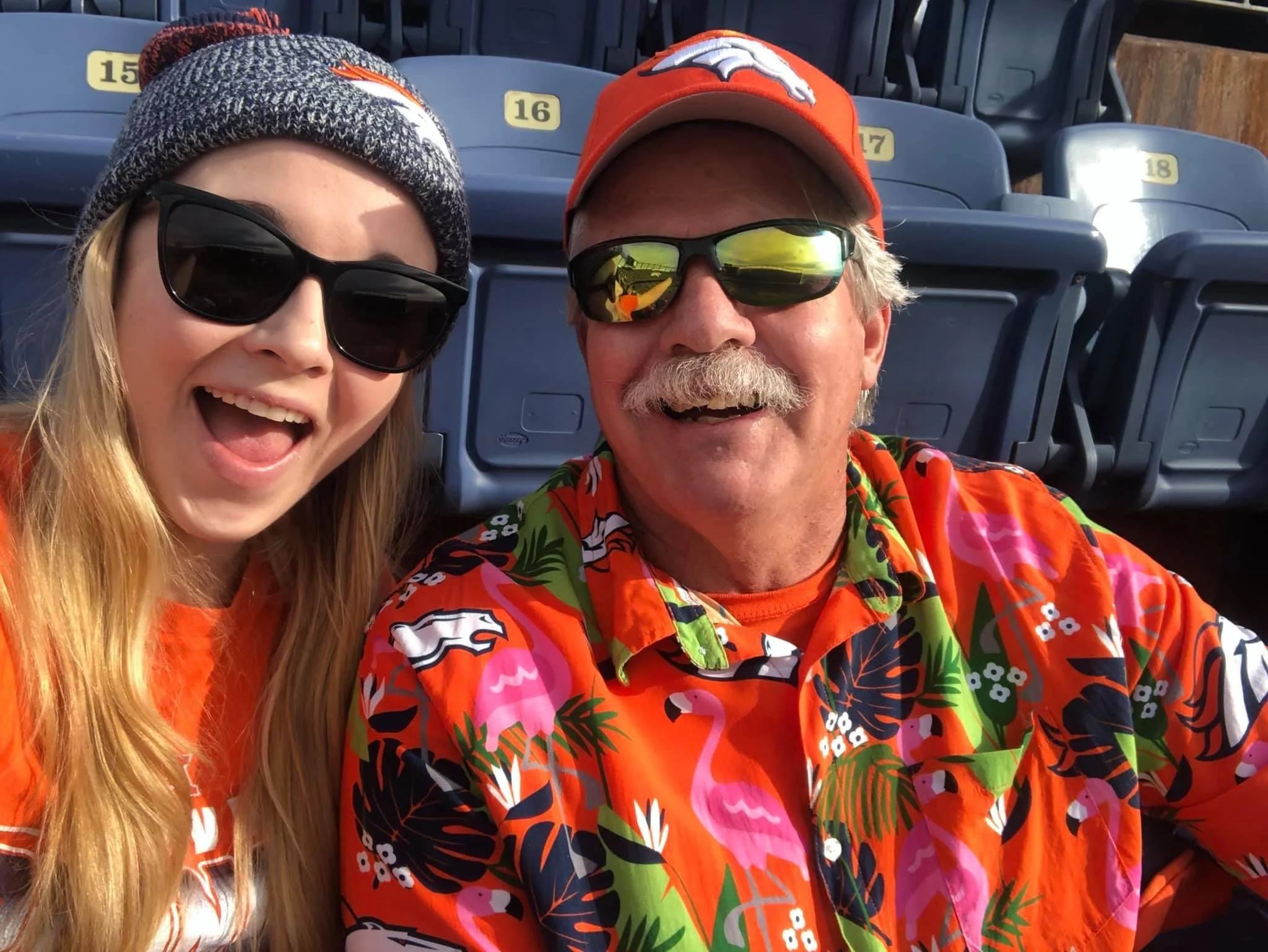 A blonde girl wearing sunglasses and a beanie smiles next to an older man wearing a denver broncos hat, sunglasses, and an orange Broncos hawaiian shirt