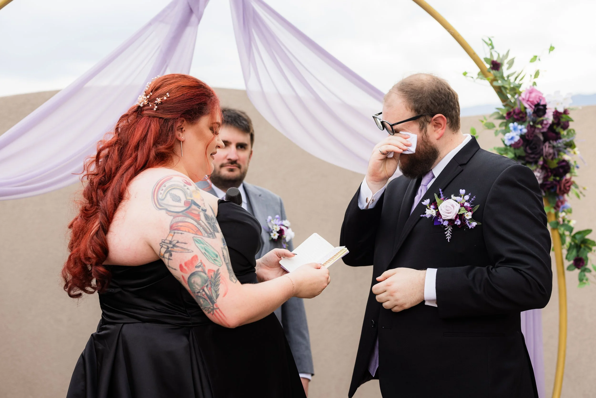 The officiant looks on as a tattooed bride reads her vows to a groom who is wiping away tears during their wedding ceremony, planned by JayBird Weddings and Events.