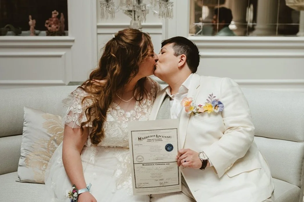 Two brides hold up their Colorado marriage license and kiss on their wedding day, planned by JayBird Weddings and Events