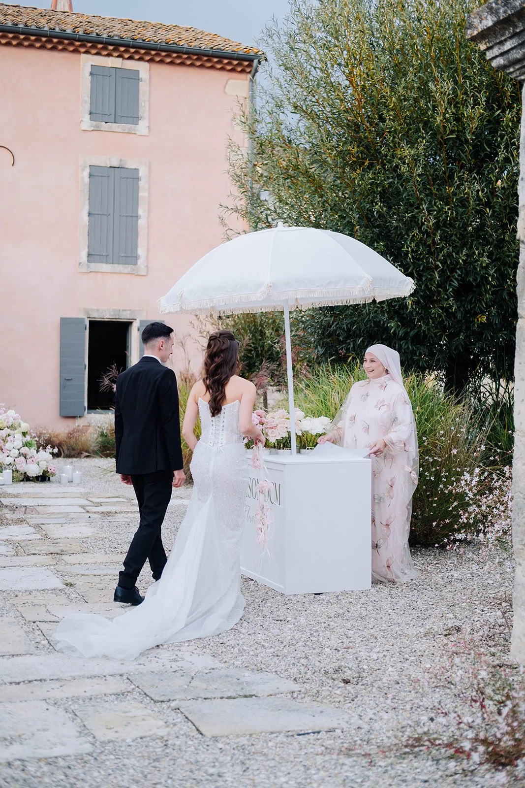 Un mariage avec un couple en tenue de mariée et de marié, échangeant des vœux sous un parasol blanc, avec une femme en hijab célébrant la cérémonie, dans un jardin fleuri.