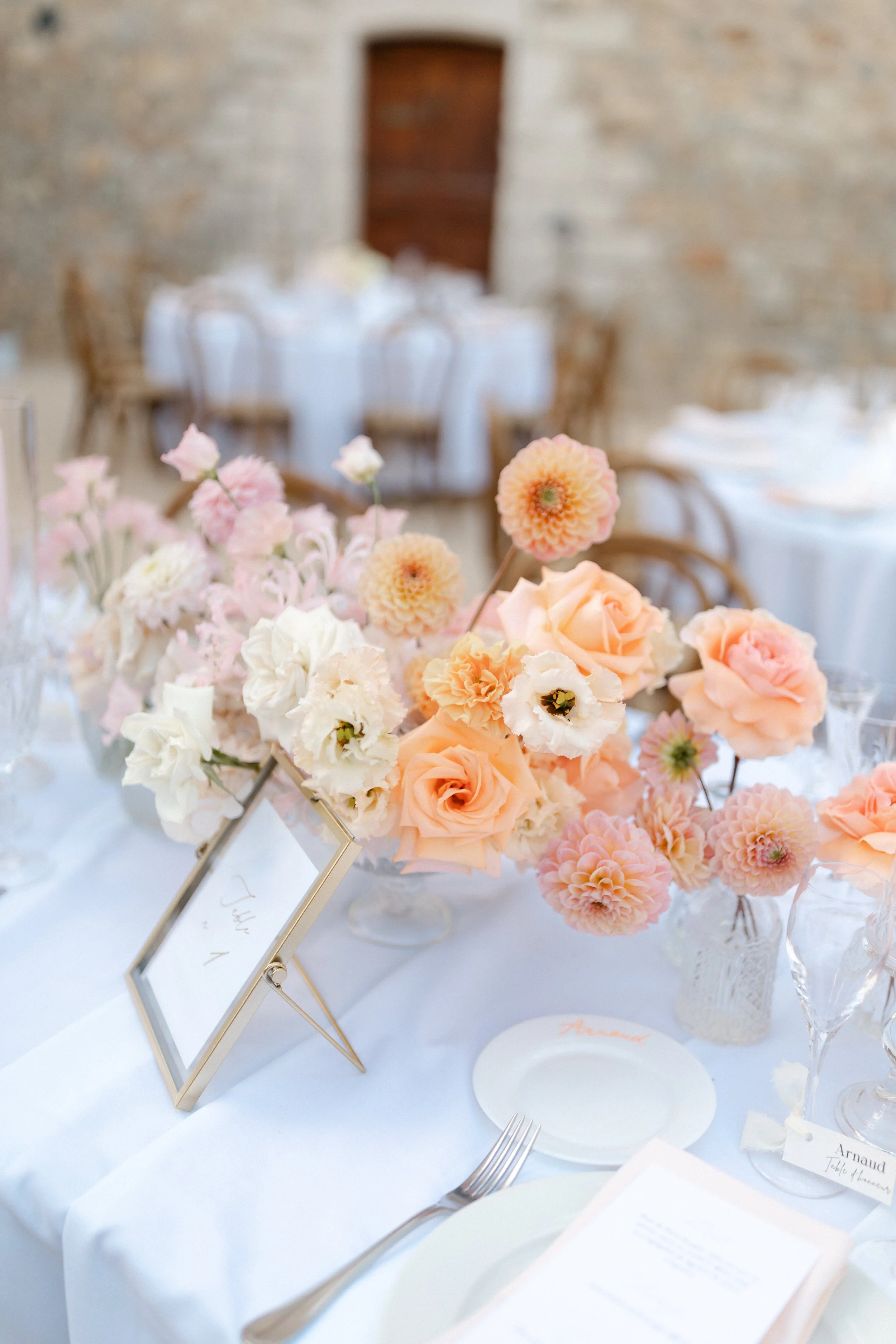 Centre de table avec un bouquet de fleurs pastel dans un vase en verre, sur une table de mariage décorée avec une nappe blanche, assiettes, couverts, et un cadre avec un menu. En arrière-plan, une salle avec des murs en pierre et des tables avec une 