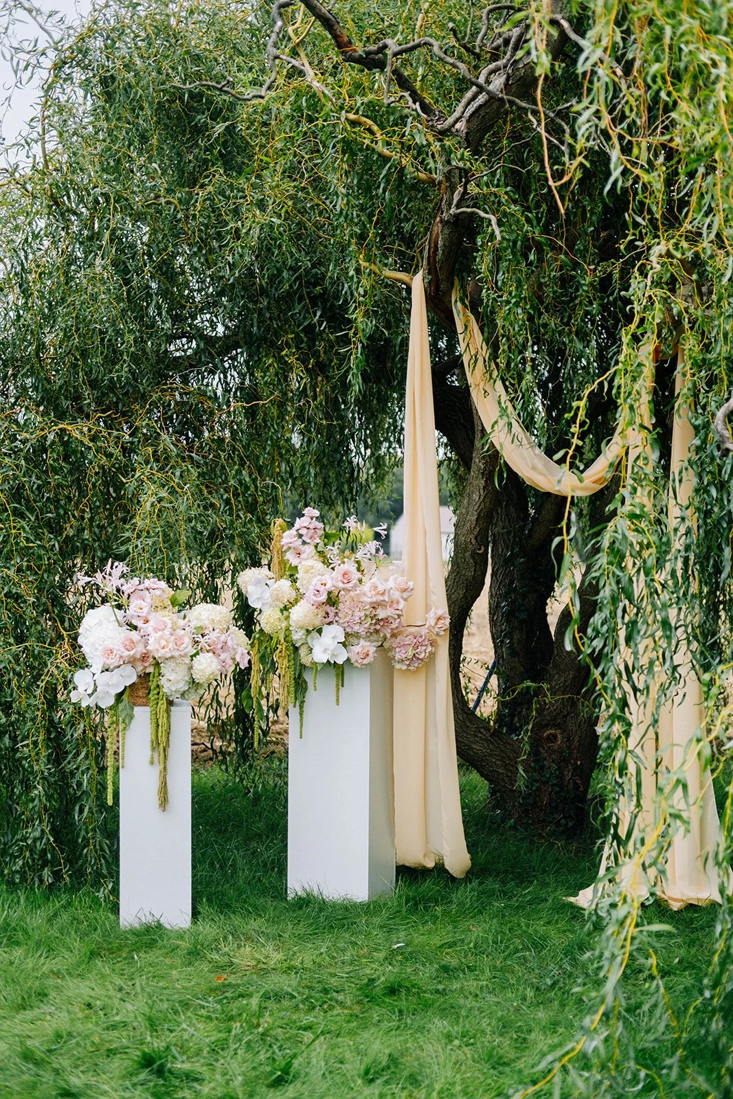 Décor de mariage avec des arrangements floraux pastel sous un arbre, avec des voiles en tissu beige accroché à ses branches.