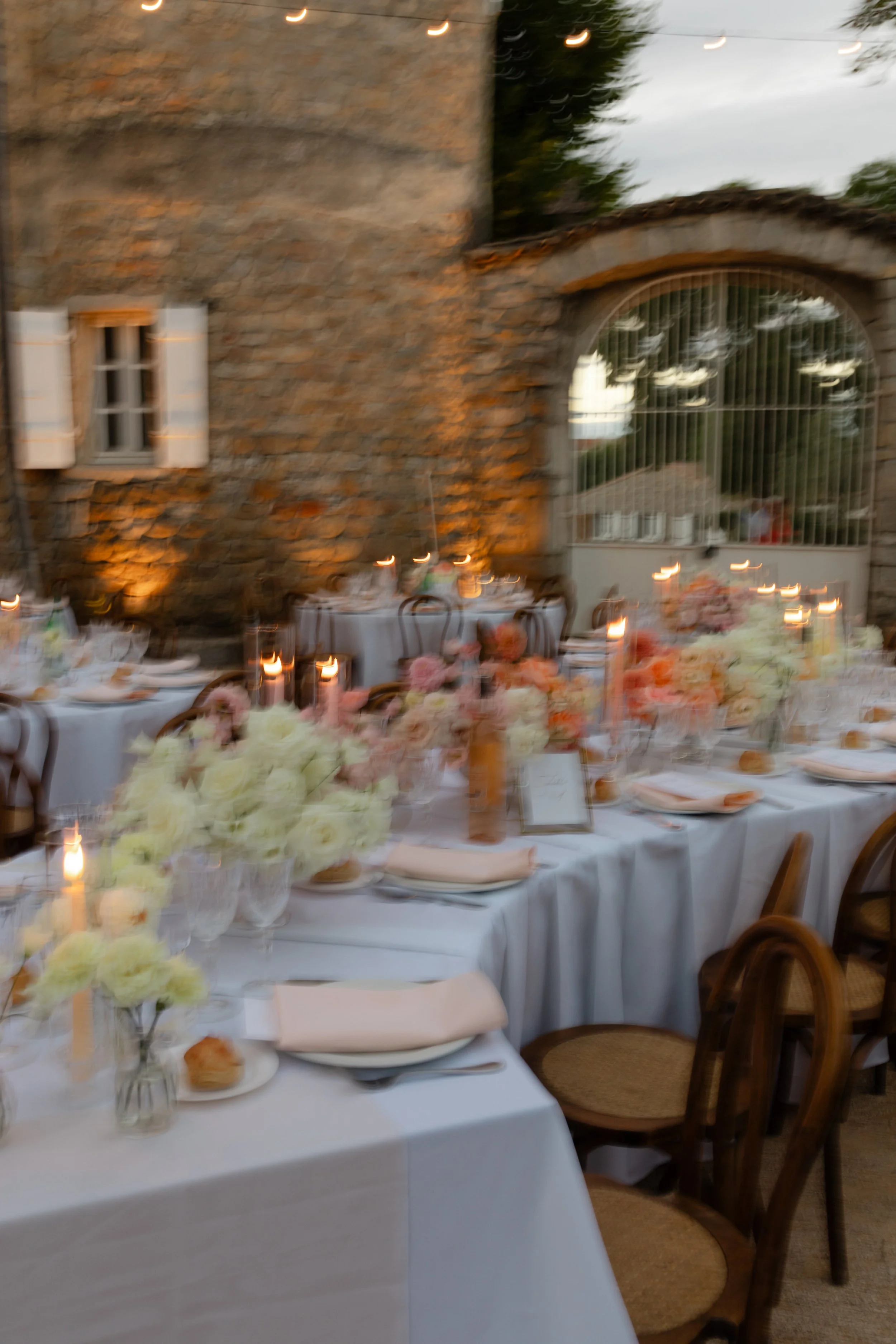 Une table de banquet décorée avec des fleurs blanches, des bougies et des assiettes, dans un style rustique avec un mur en pierre et une grande porte en fer, probablement lors d'une célébration ou d'un événement en extérieur.