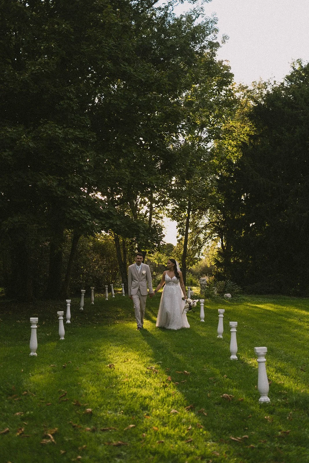 Un couple de mariés marche main dans la main dans un jardin ensoleillé, entouré d'arbres et de pelouse verte, avec des bornes blanches décoratives le long de leur chemin.