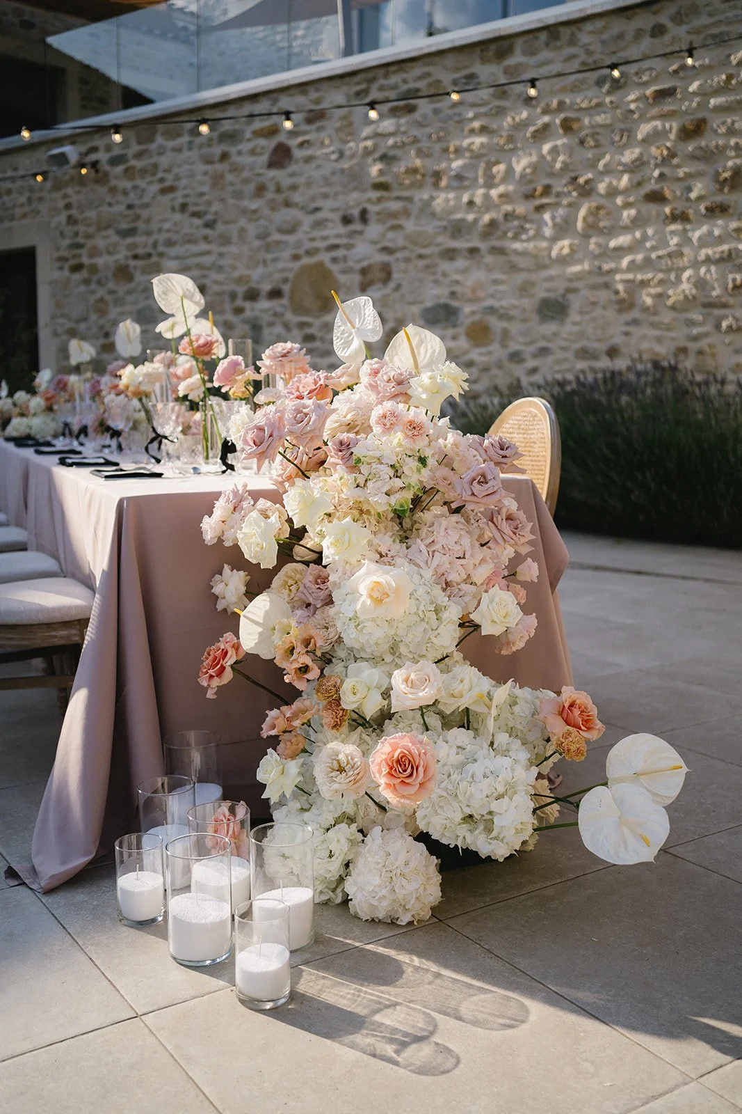 Table décorée avec un arrangement floral rose et blanc, bougies dans des verres à côté, dans un cadre en plein air avec un mur en pierre