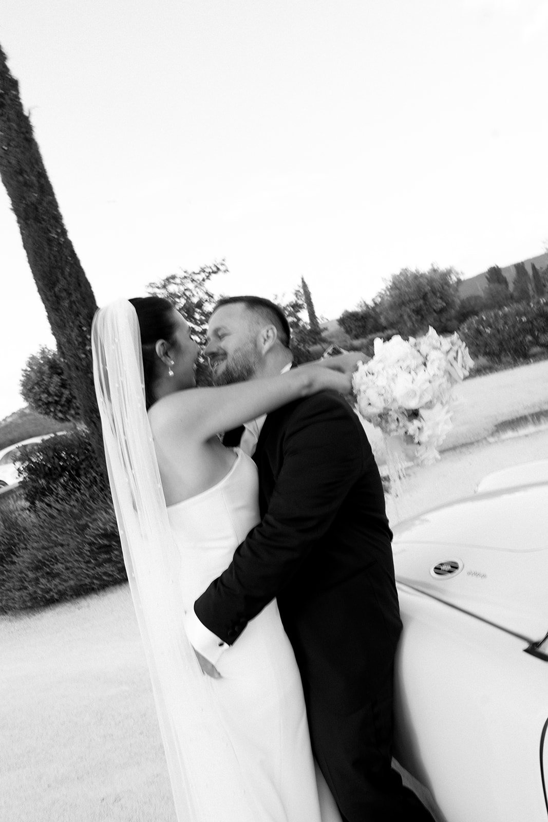 Un couple de mariés s'embrassant avec une voiture décorée et un bouquet de fleurs dans le contexte d'un mariage en extérieur.