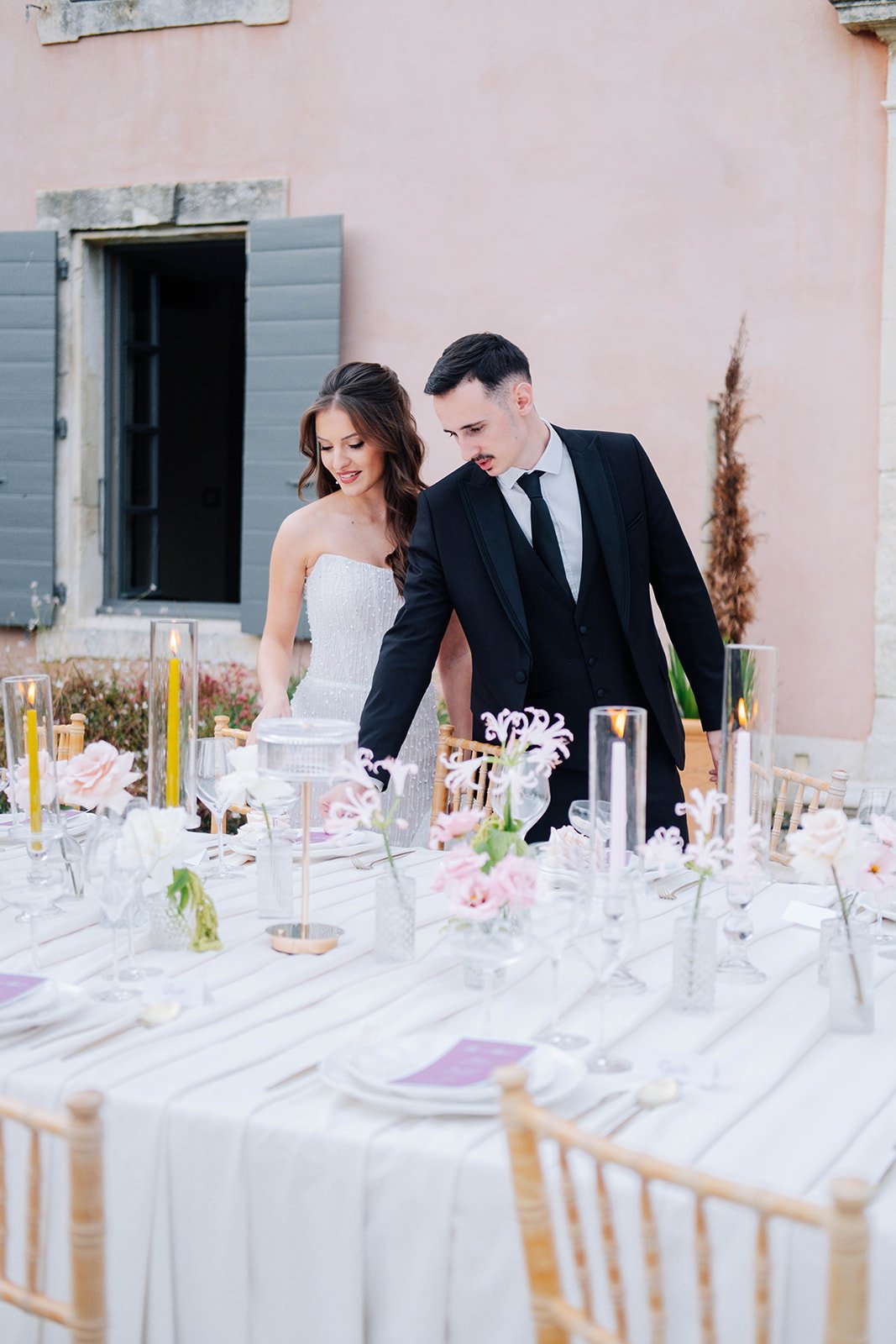 Un couple en costume de mariage, la femme en robe blanche et l'homme en costume noir, se tient près d'une table de mariage décorée avec des fleurs et des bougies, dans une cour extérieure avec un mur en pierre et une fenêtre ouverte.