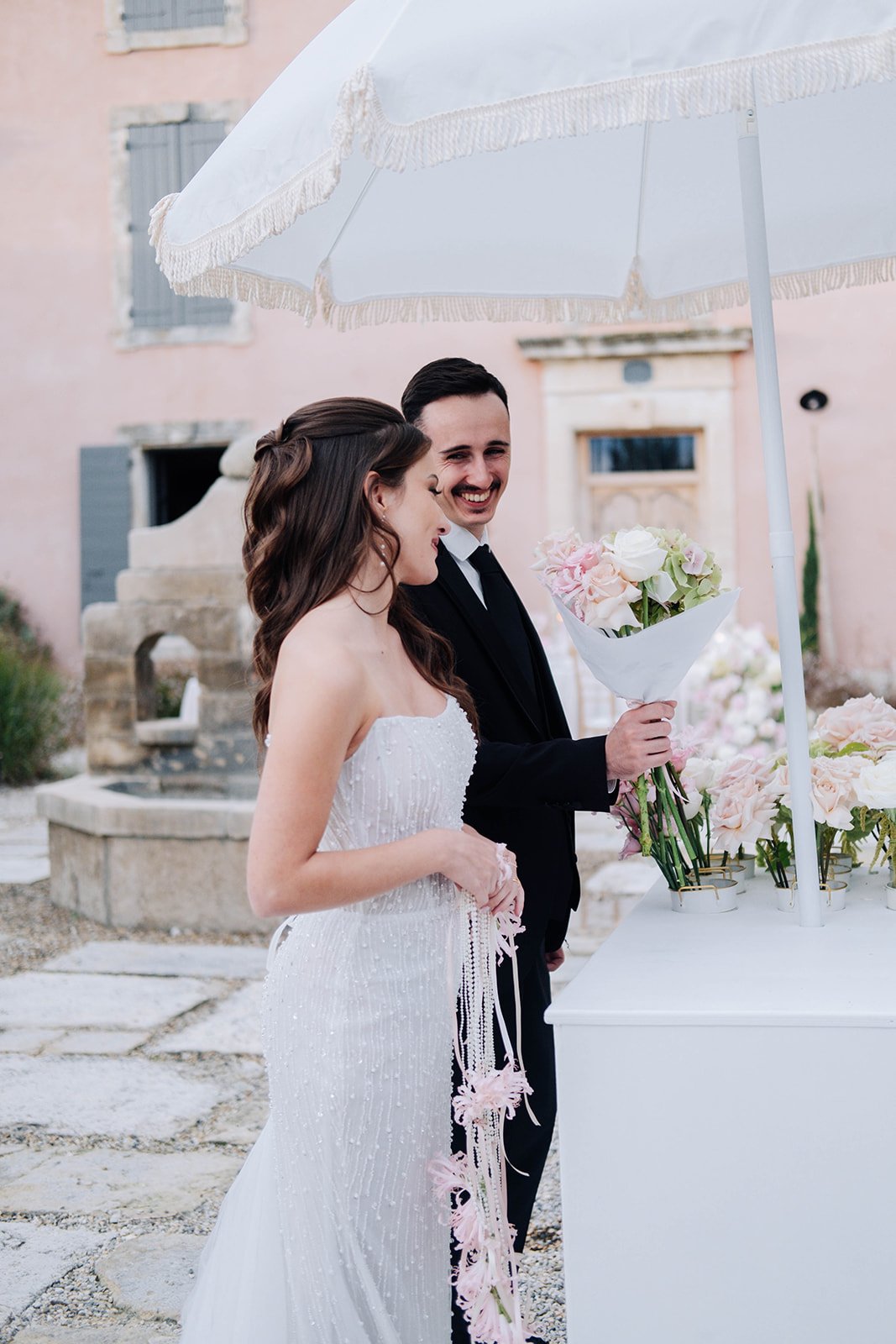 Un couple en costume de mariage devant une fontaine, échangeant un sourire, sous un parasol blanc avec bordures en pompons, avec des fleurs dans une enveloppe et d'autres fleurs devant eux.