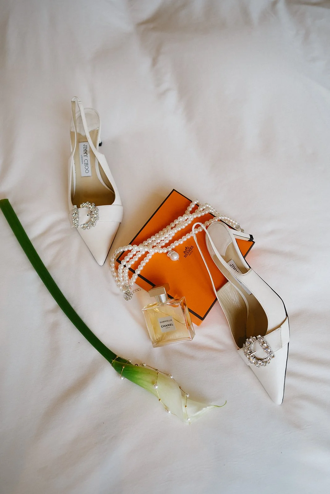 Une paire de chaussures blanches de marque Jimmy Choo avec des détails en cristal, une boîte orange Hermès, des colliers de perles, un flacon de parfum Chanel Gabrielle, une orchidée blanche, et un bracelet orné de perles, tous disposés sur un lit bl