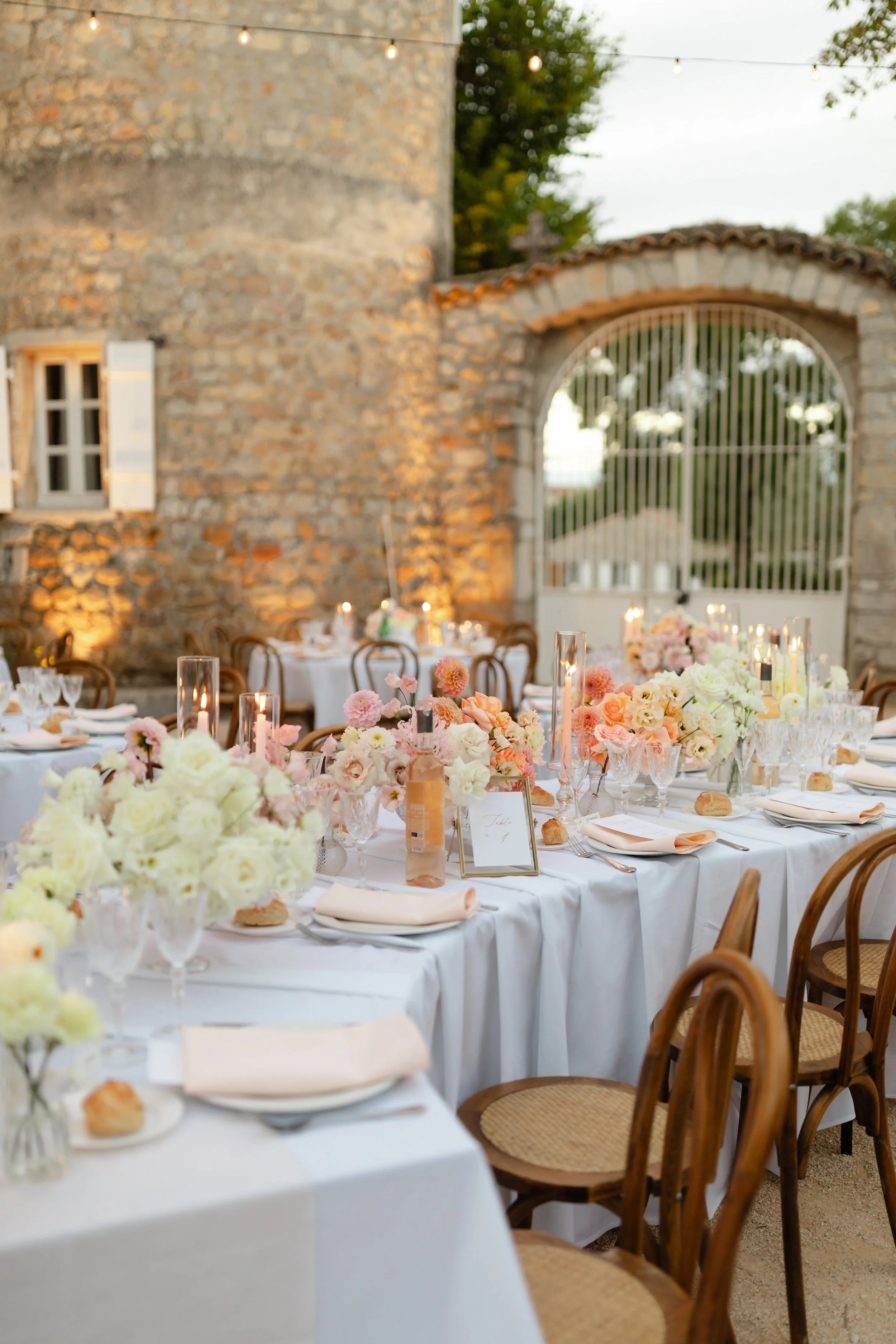 Décor de réception en plein air avec tables rondes ornées de fleurs pastel, bougies et vaisselle élégante, dans un cadre rustique avec un mur en pierre et une porte en fer forgé.