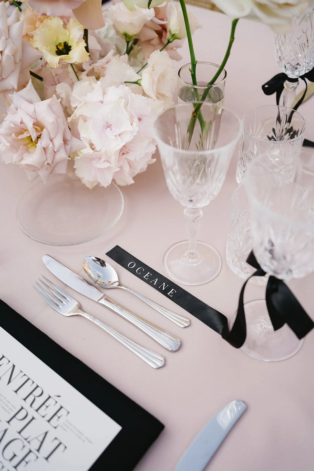 Un arrangement floral avec des fleurs blanches et roses dans des vases en cristal, une assiette, une fourchette, un couteau et une cuillère en argent, des verres en cristal et un menu de table avec une étiquette 