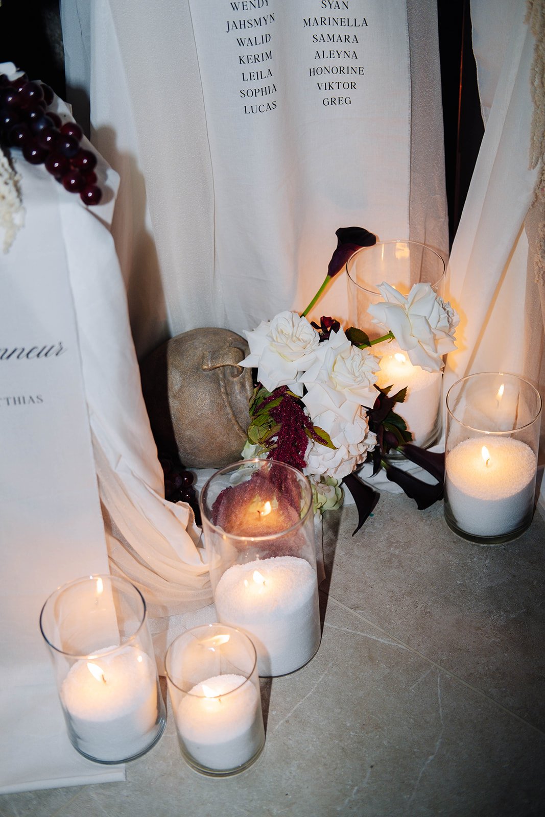 Décoration funéraire avec bougies blanches dans des verres, fleurs blanches, une pierre peinte, un panneau blanc avec des noms en noir.