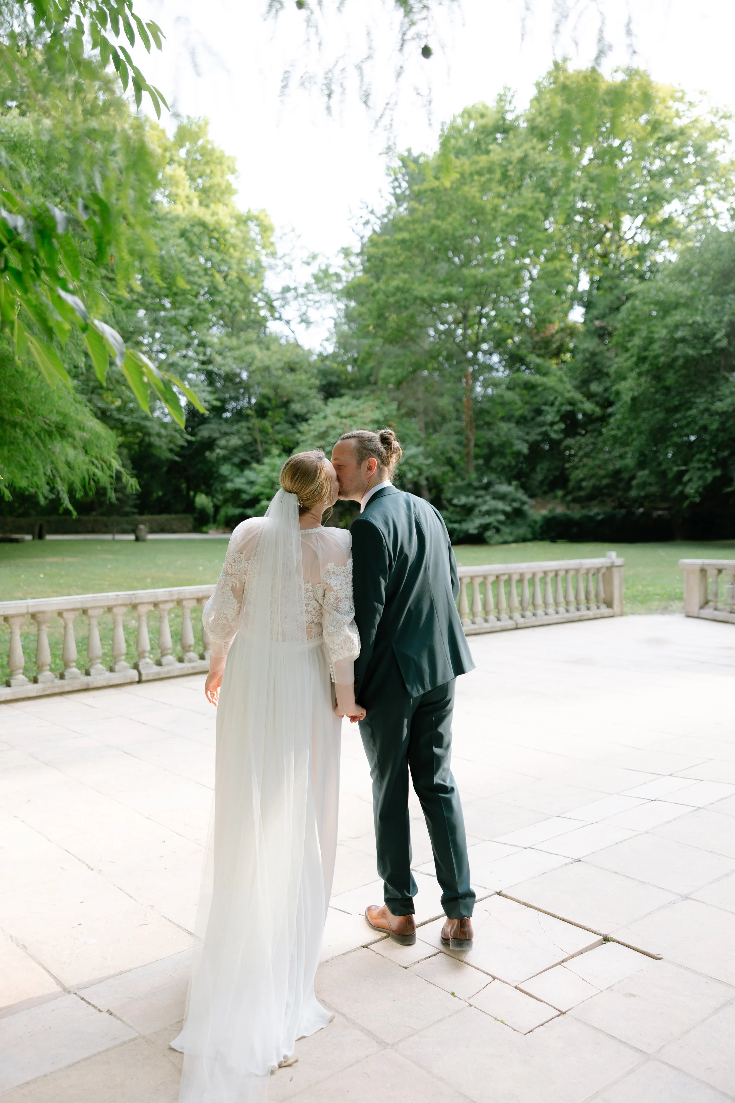 Un couple de mariés s'embrassant dans un parc verdoyant, avec des arbres en arrière-plan et un sol en pierre, lors d'une cérémonie de mariage.
