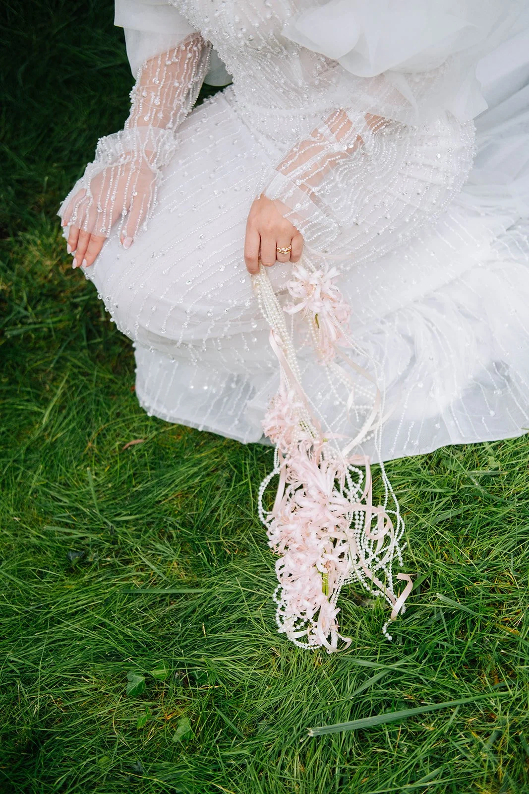 Zoom sur une robe de mariée blanche ornée de perles, portée par une personne assise sur de l'herbe verte, tenant un bouquet de fleurs rose pâle avec des rubans et des perles.