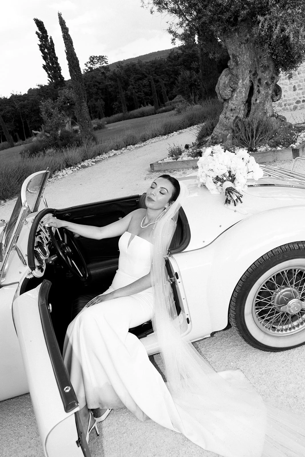 Une femme en robe de mariée assise dans une voiture de mariage avec un bouquet de fleurs sur le capot, dans un jardin avec des arbres et des arbustes.