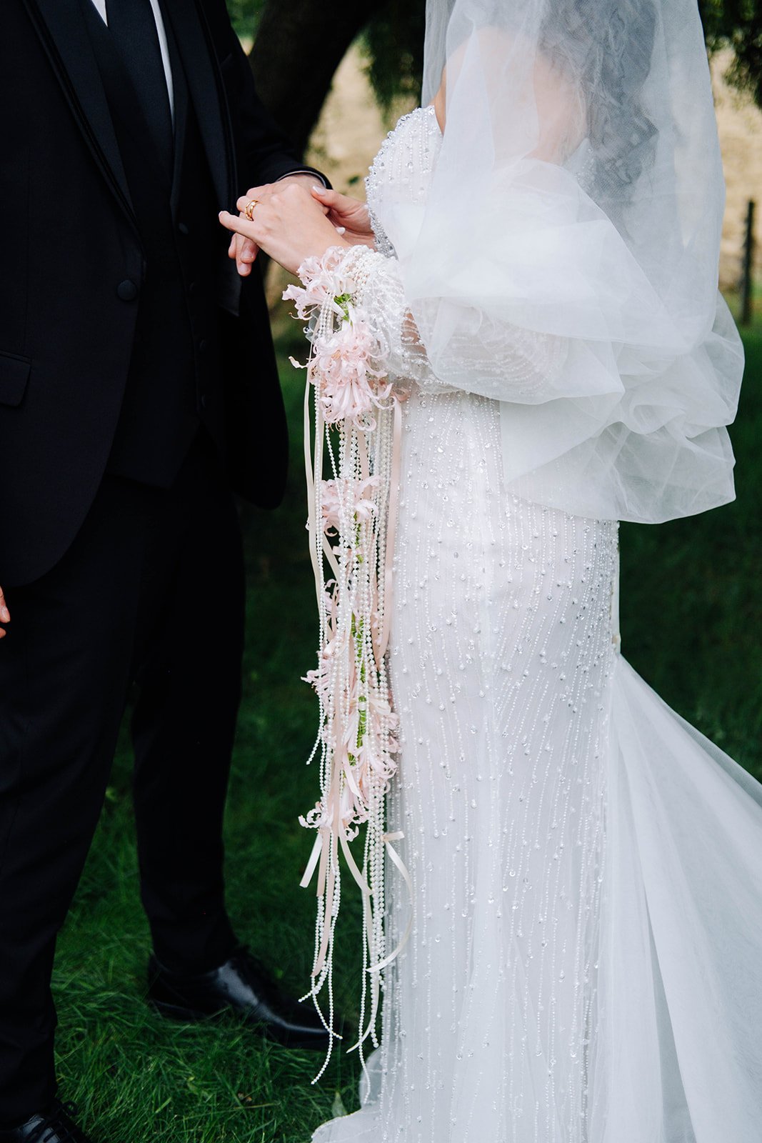 Un couple se tenant la main lors d'un mariage en plein air. La mariée porte une robe blanche ornée de perles et une voile, tenant un bouquet de fleurs roses. L'homme porte un costume noir.