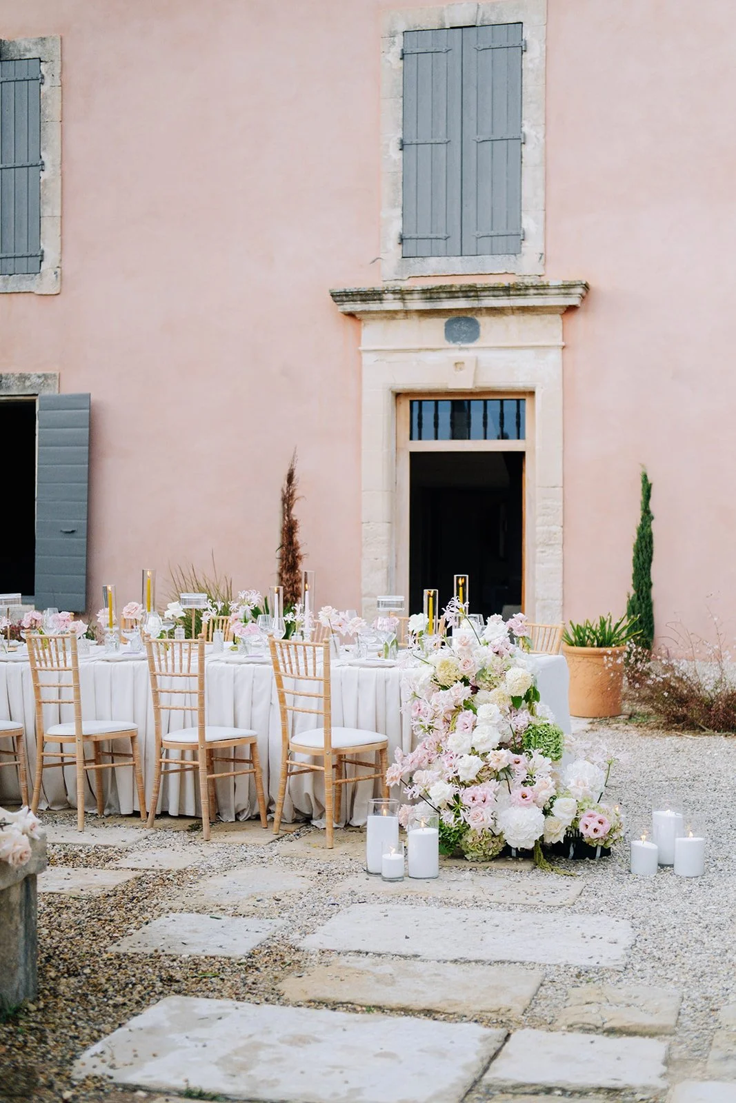 Décor de mariage avec une table ronde, chaises en bois clair, fleurs pastel et bougies dans une cour pavée, devant un bâtiment en pierre rose avec volets bleus.