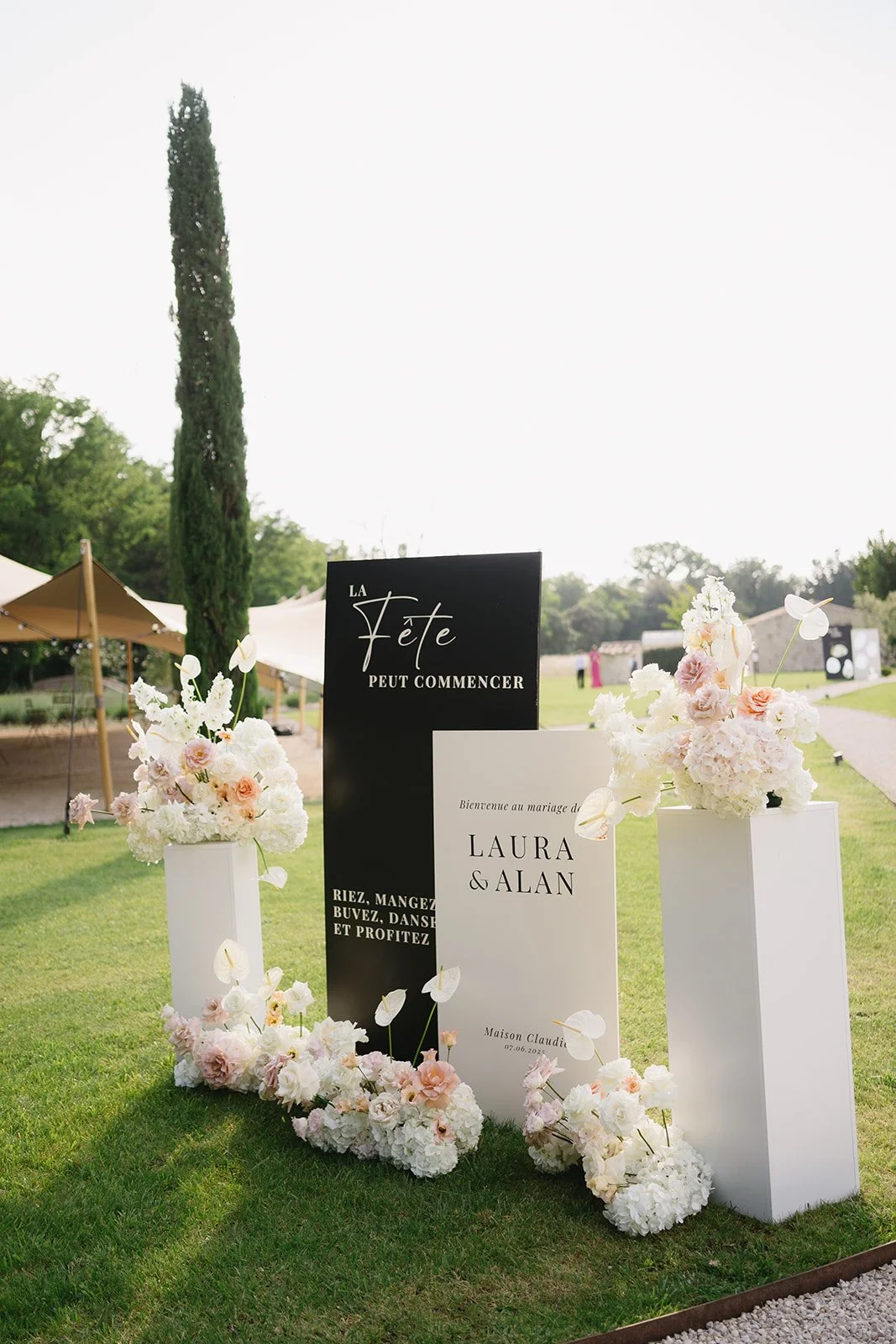 Décor de mariage avec deux arrangements floraux blancs et roses, panneaux d'accueil en français pour Laura et Alan, et un fond extérieur avec tentes, arbres et ciel clair.