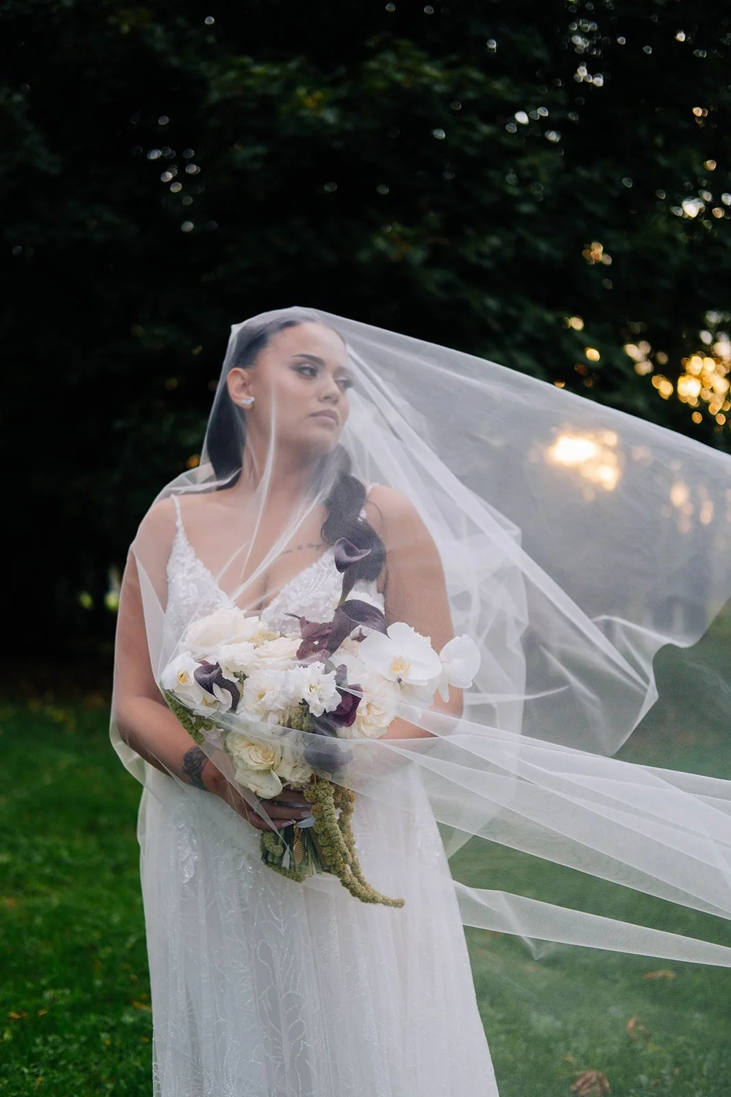 Une mariée avec une robe blanche, tenant un bouquet de fleurs, portant un voile, en extérieur avec des arbres en arrière-plan.