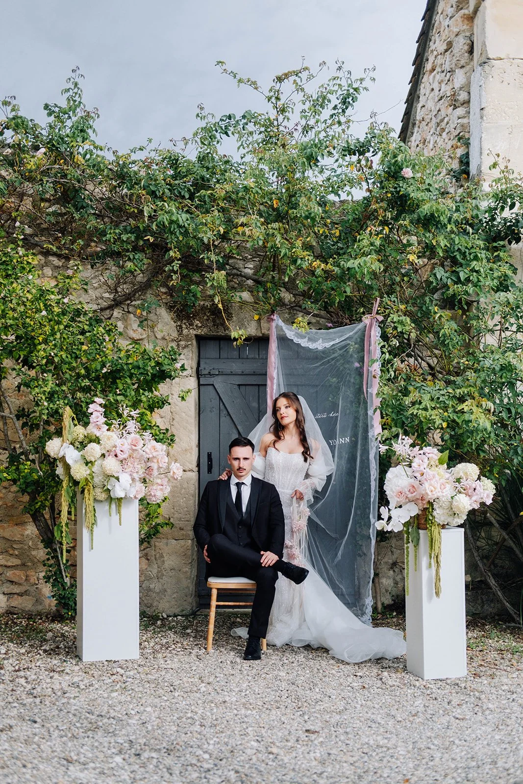 Un couple en robe de mariée et costume, posant dans un décor floral avec des bouquets de fleurs roses et blanches sur des colonnes, devant un mur en pierre avec une porte en bois gris.