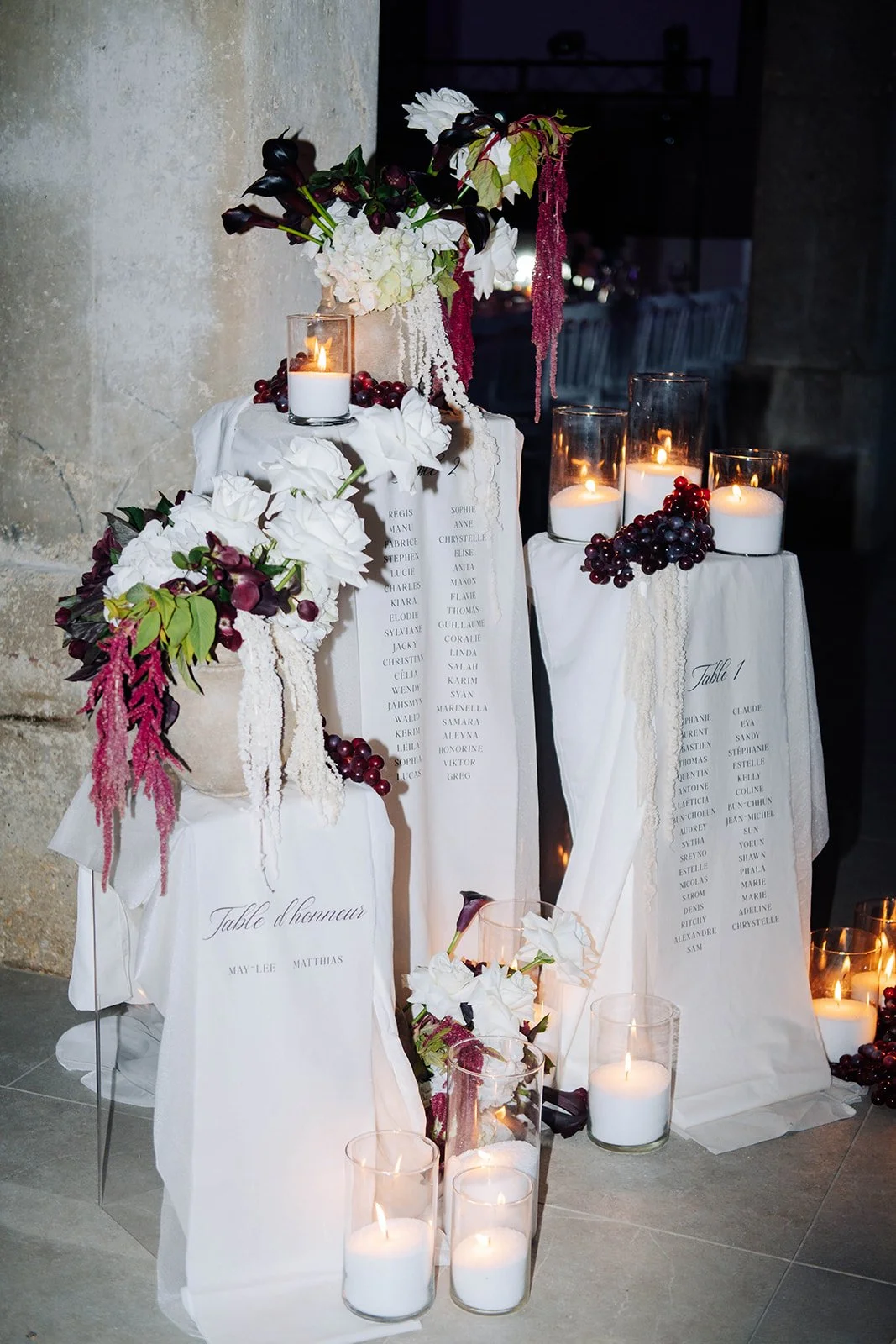Décoration de cercueil de mariage avec fleurs blanches et pourpres, bougies dans des verres, et listes de noms en français.