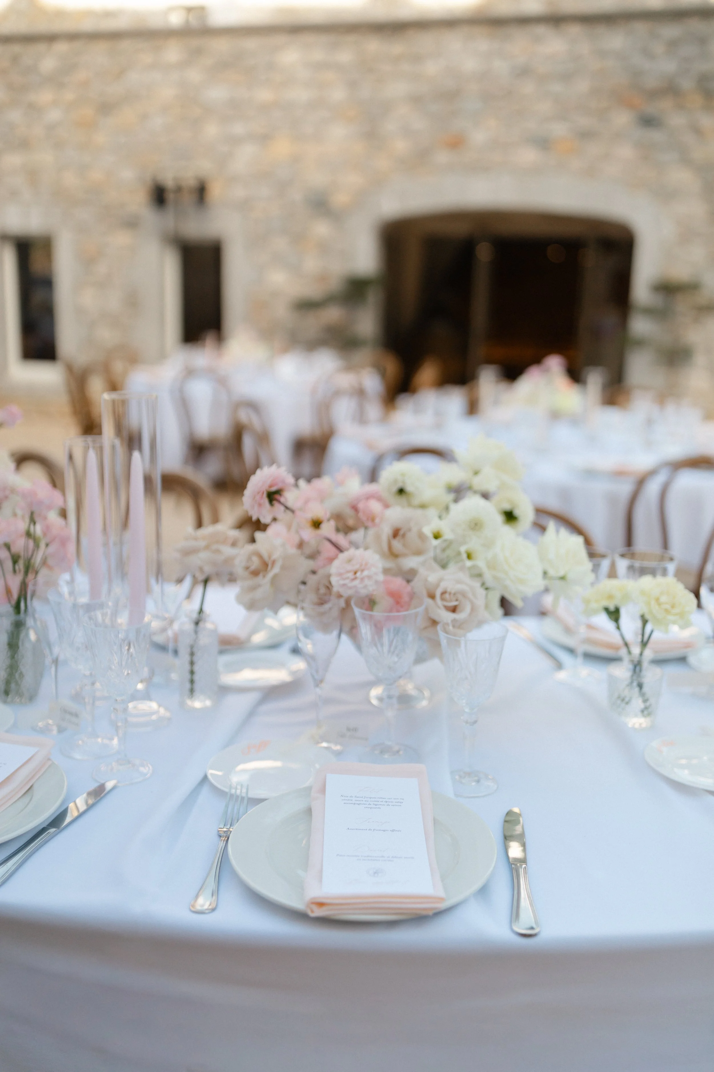 Table de banquet avec des fleurs blanches et roses, des verres à vin, des assiettes, des couverts, et des menus en papeterie, décorée pour un événement formel, dans une salle avec mur en pierre et cheminée.