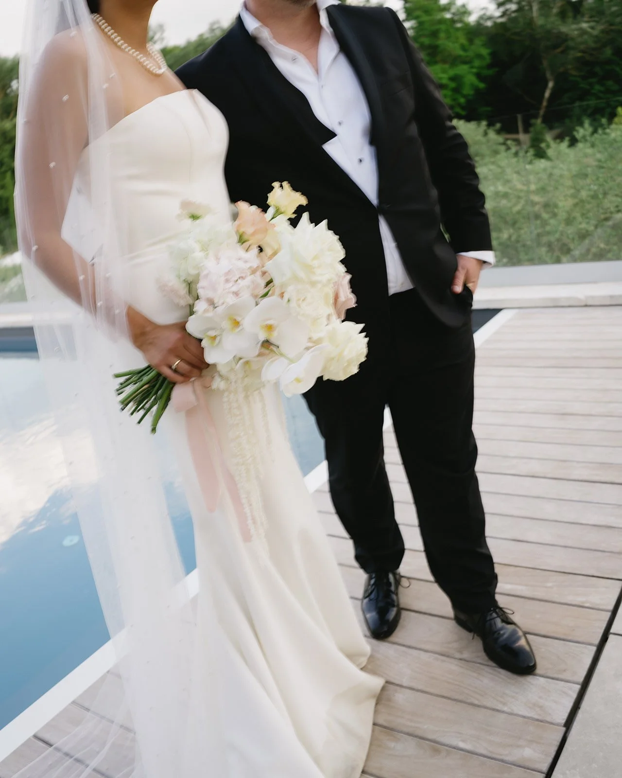Un couple de mariés posant pour une photo, la mariée tenant un bouquet de fleurs blanches, l'homme en costume noir avec chemise blanche, en extérieur.