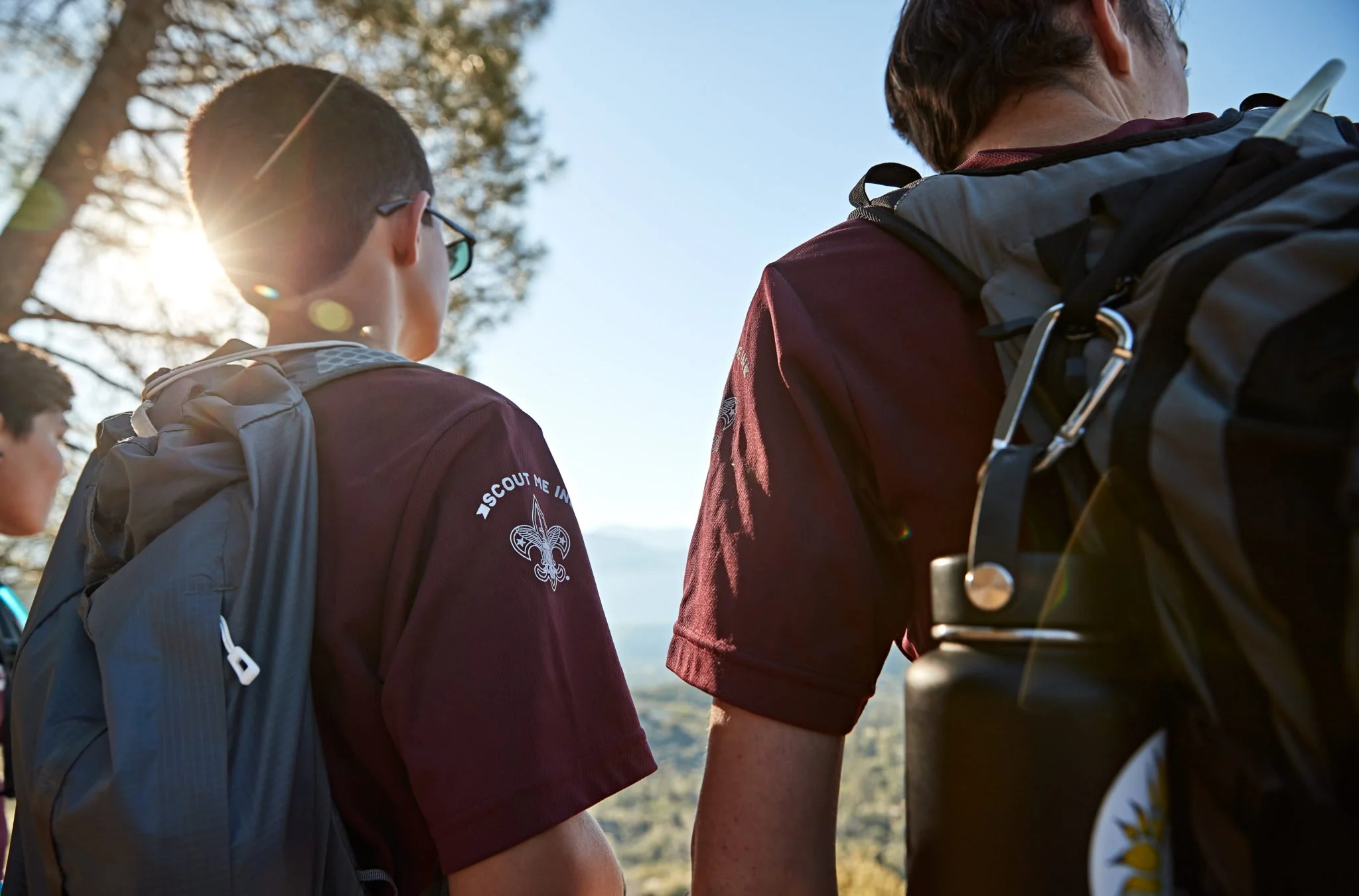 Two Boy Scouts wearing maroon scout uniforms with backpacks standing outdoors on a sunny day, with trees and a landscape in the background.