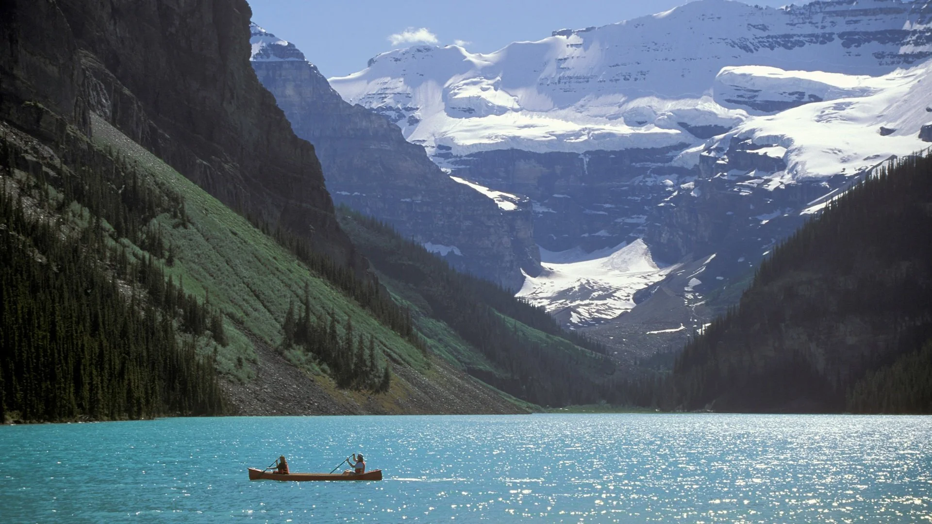 A scenic mountain lake with turquoise waters, surrounded by green forested slopes and snow-capped peaks in the background, with a small boat carrying two people on the water.